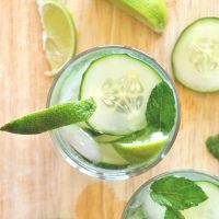 Glasses of our Cucumber Cooler Cocktail recipe topped with cucumbers slices, mint leaves, and limes