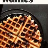 A lemon blueberry waffle cooking in a waffle iron with text above it that says vegan and gluten-free