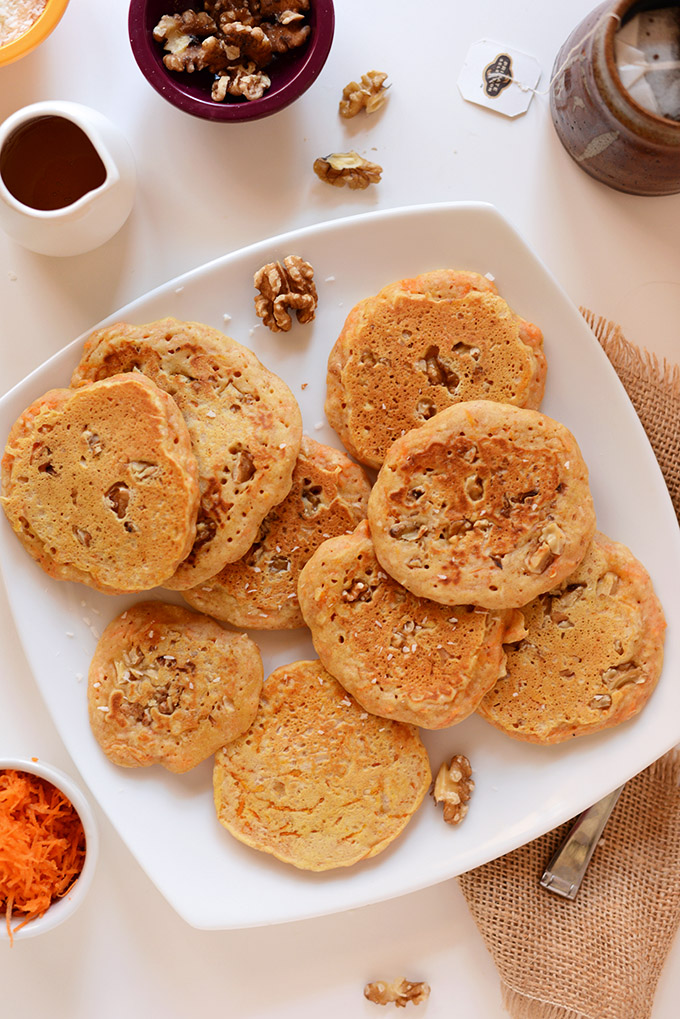 Large plate of freshly cooked Carrot Coconut Pancakes alongside syrup and walnuts