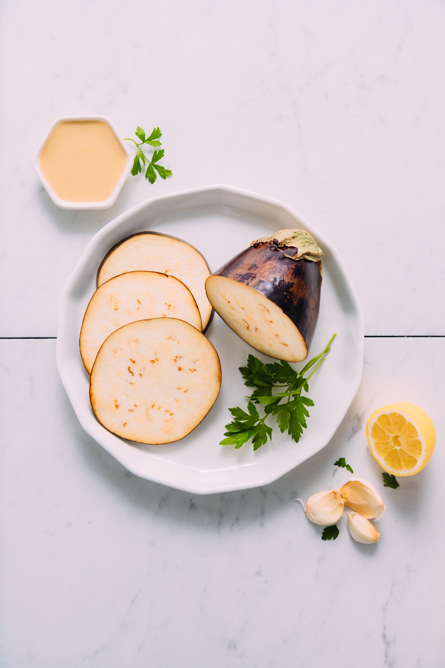 Partially sliced eggplant next to parsley, garlic, and lemon