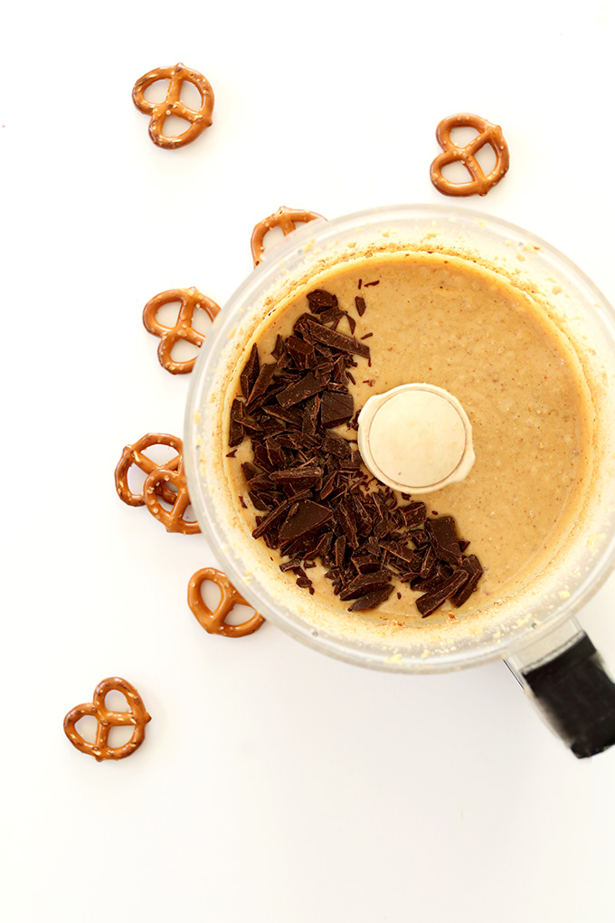 Pretzels around a food processor filled with homemade peanut butter and chopped vegan chocolate