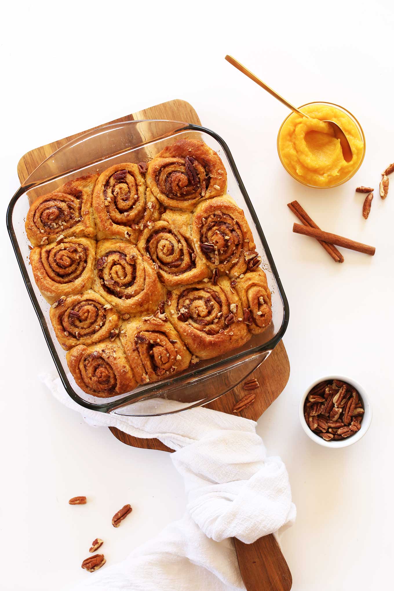 Pan of freshly baked Vegan Pumpkin Cinnamon Rolls alongside cinnamon sticks and bowls of pecans and pumpkin puree