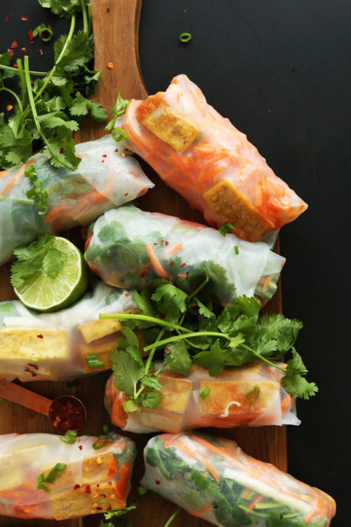 Wood cutting board with gluten-free vegan Bahn Mi Spring Rolls, dipping sauce, fresh cilantro, and lime