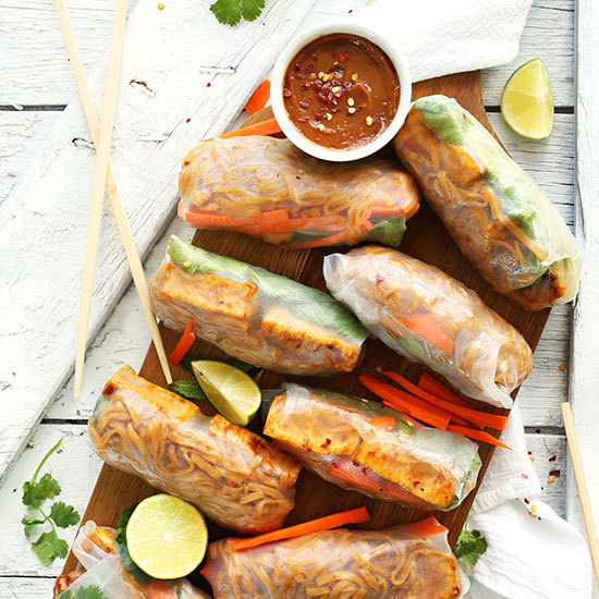 Wood cutting board filled with Pad Thai Spring Rolls, lime wedges, and a bowl of dipping sauce
