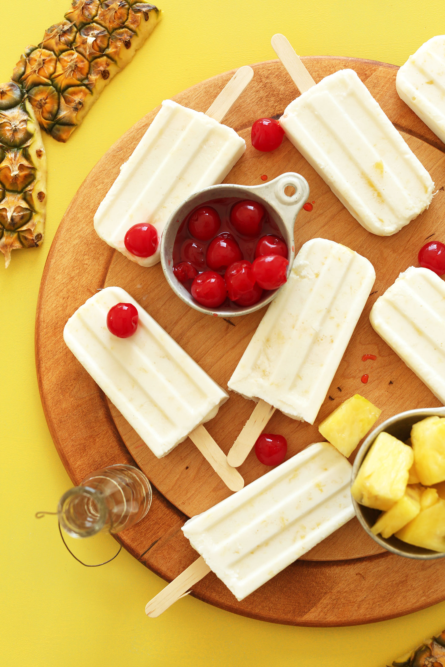 Cutting board with vegan Pina Colada Popsicles and measuring cups full of cherries and pineapple
