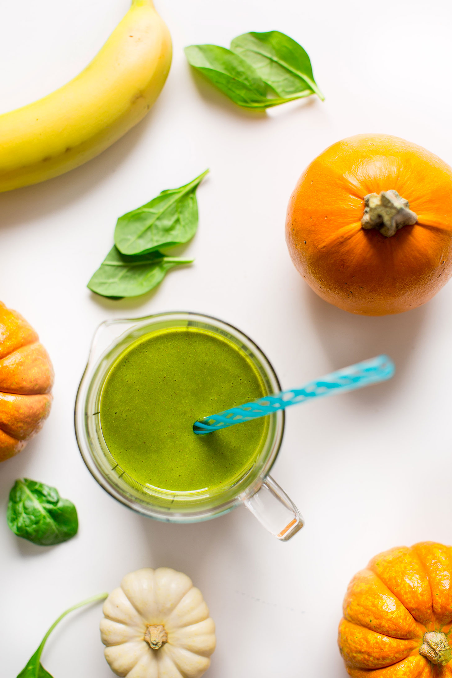 Measuring glass filled with Pumpkin Pie Green Smoothie and a straw alongside ingredients used to make it