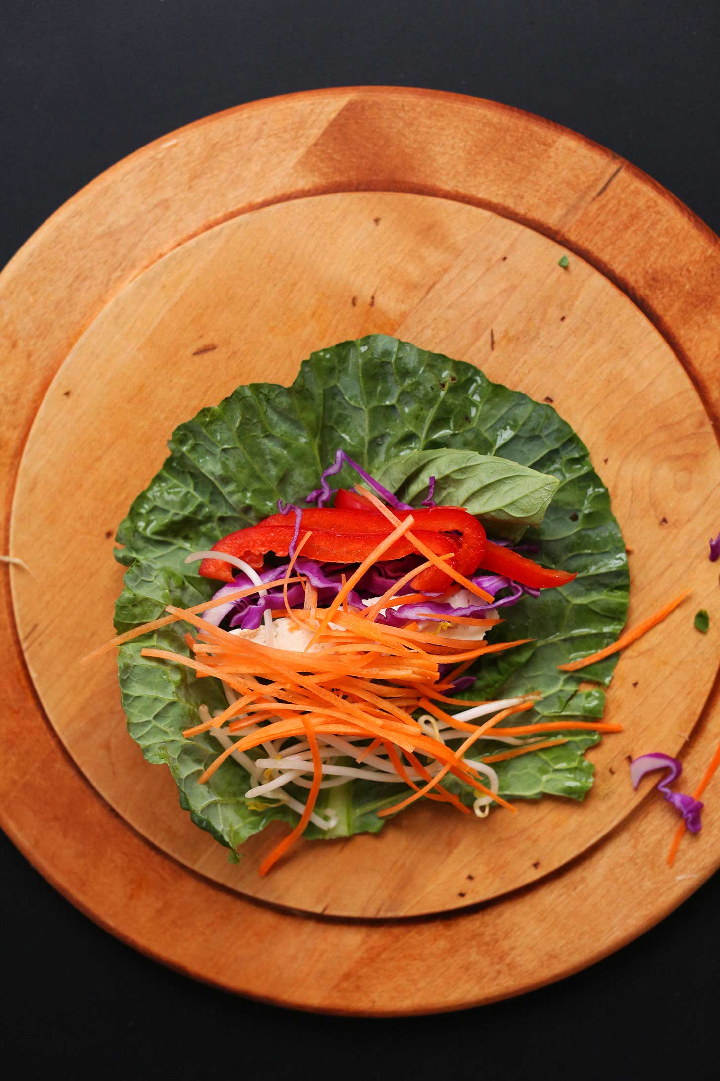 Wood cutting board with a collard green leaf and fresh ingredients for making hearty plant-based spring rolls