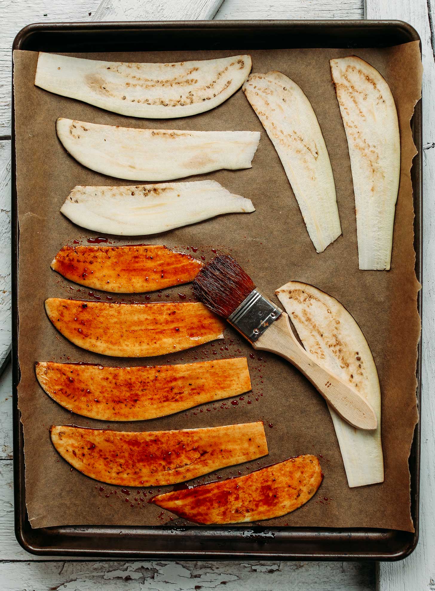 Brushing sauce onto sliced eggplant laying on a parchment-lined baking sheet