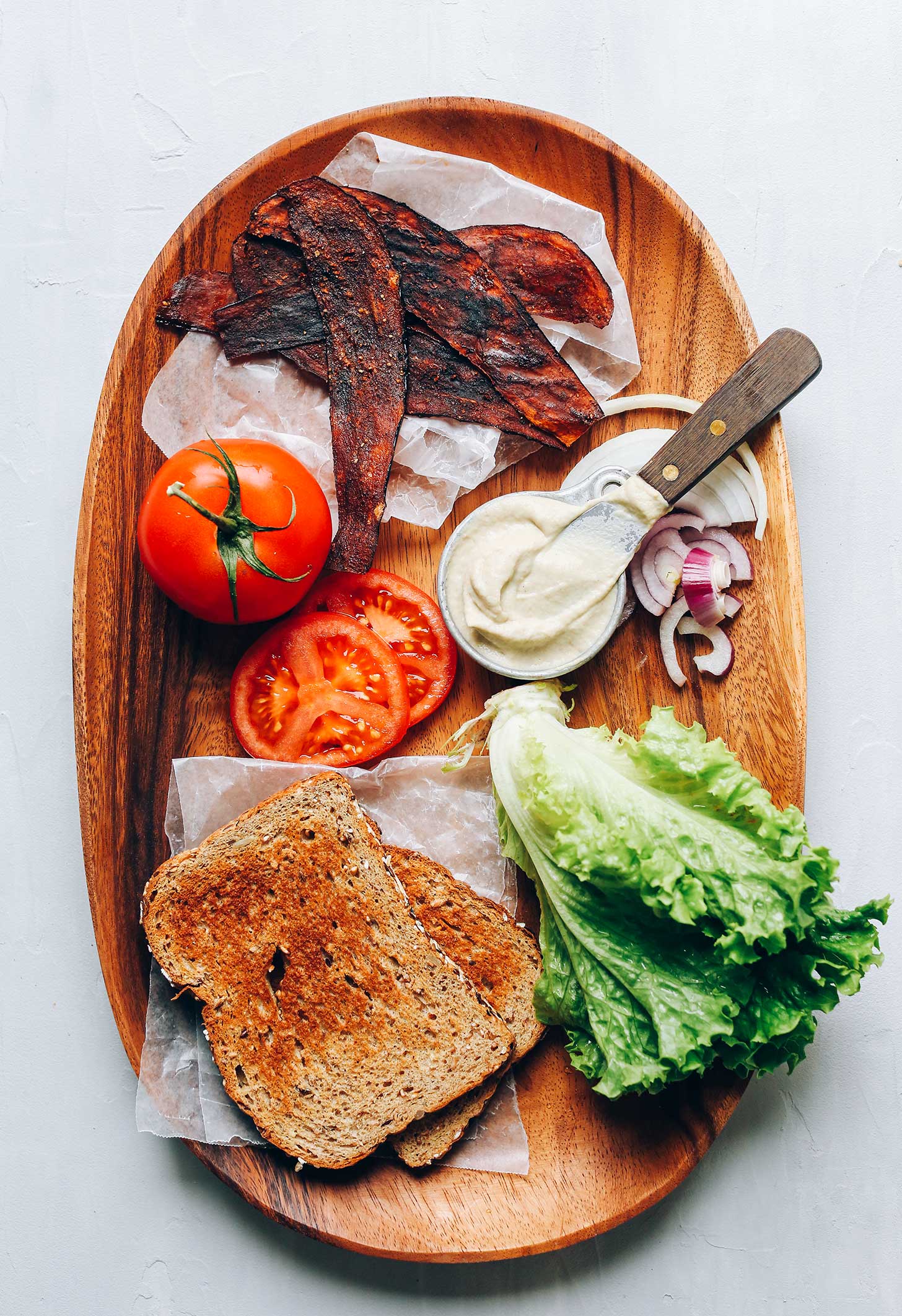 Wood platter with bread, lettuce, vegan mayo, onion, tomato, and eggplant bacon for a Vegan BLT Sandwich