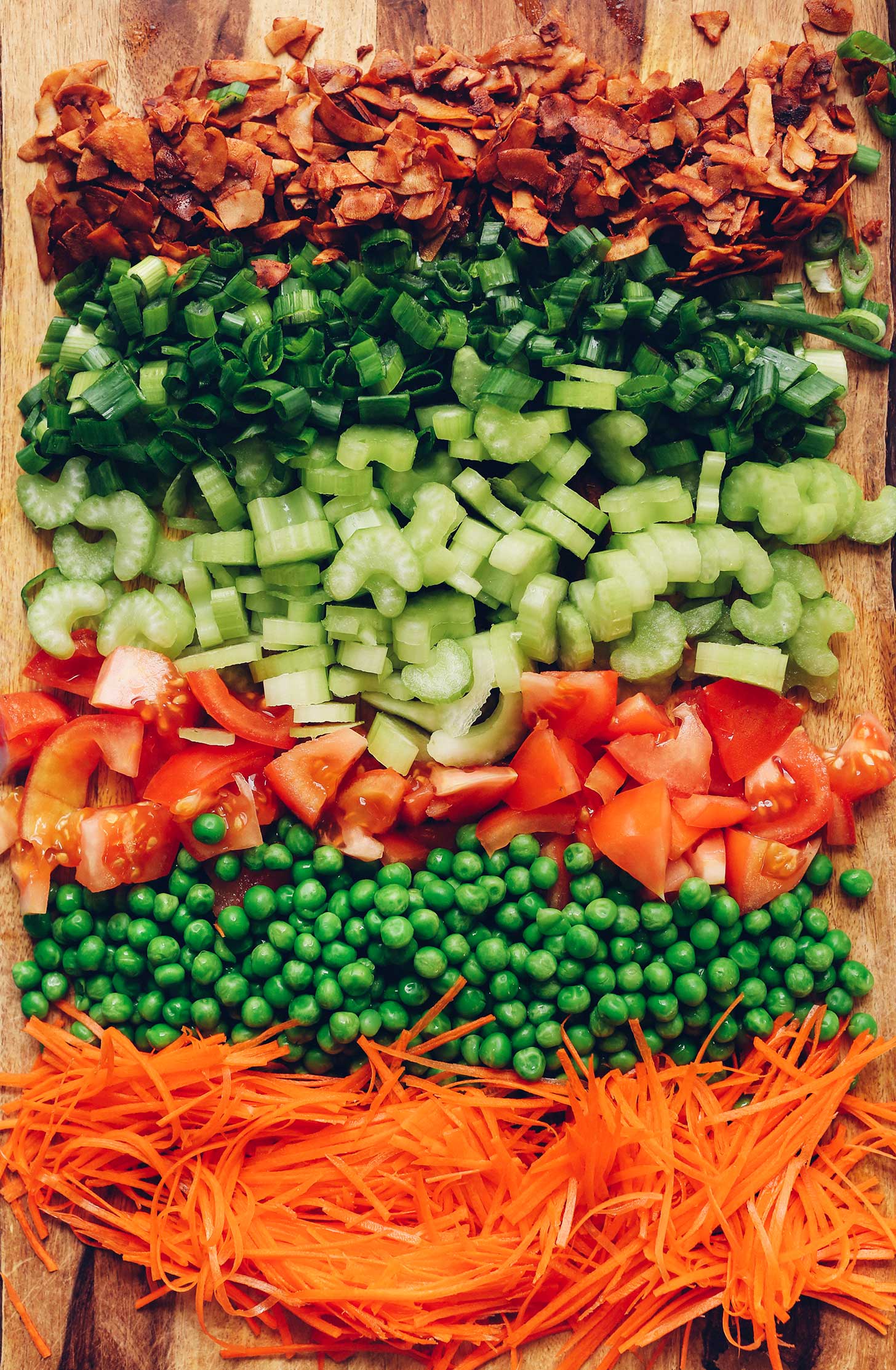 Shredded carrots, green peas, tomatoes, celery, green onion, and coconut bacon on a wood cutting board to be made into Vegan 7-Layer Salad