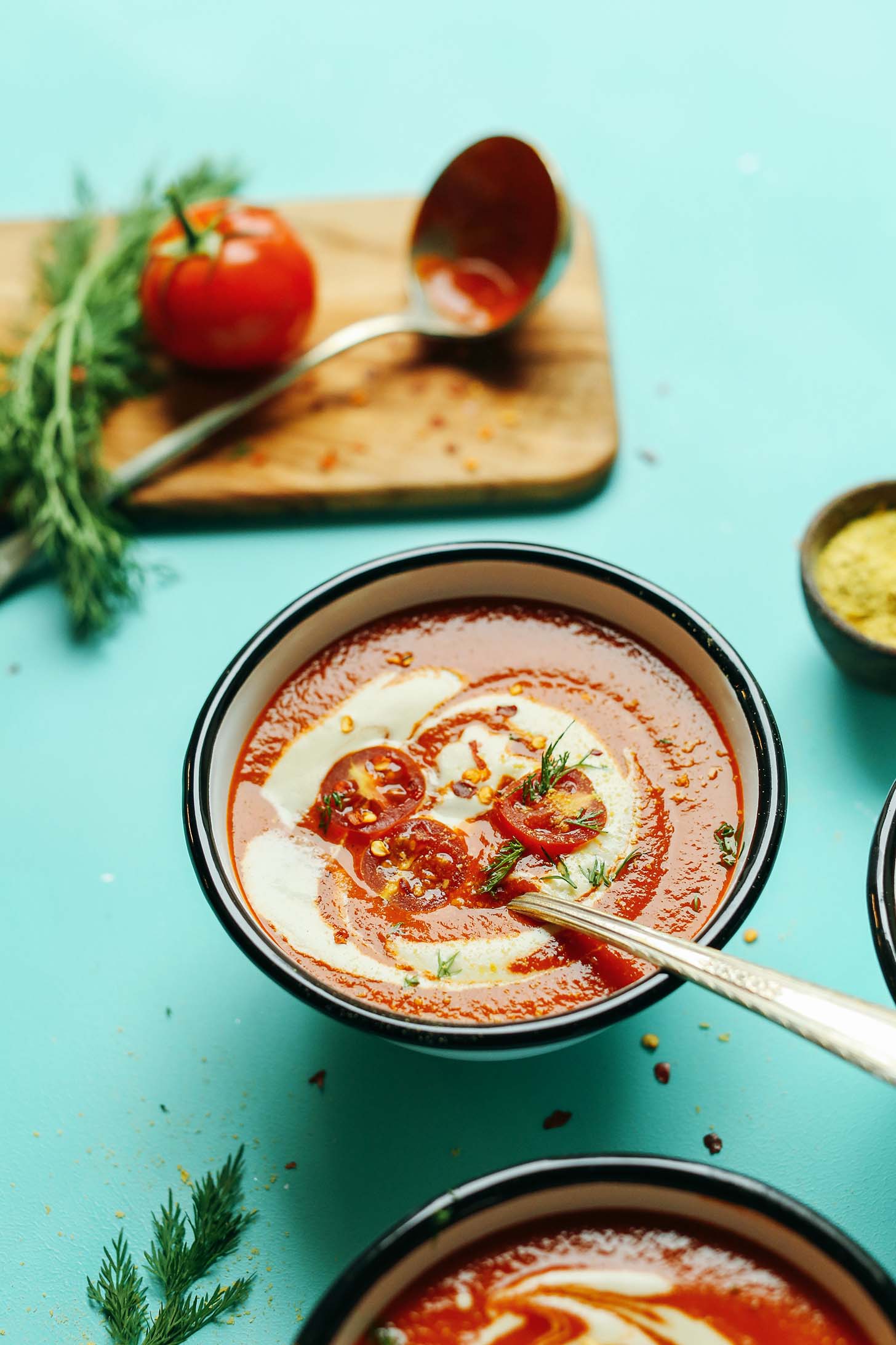 A bowl of Creamy Roasted Red Pepper Tomato Soup and a cutting board with a fresh tomato and dill
