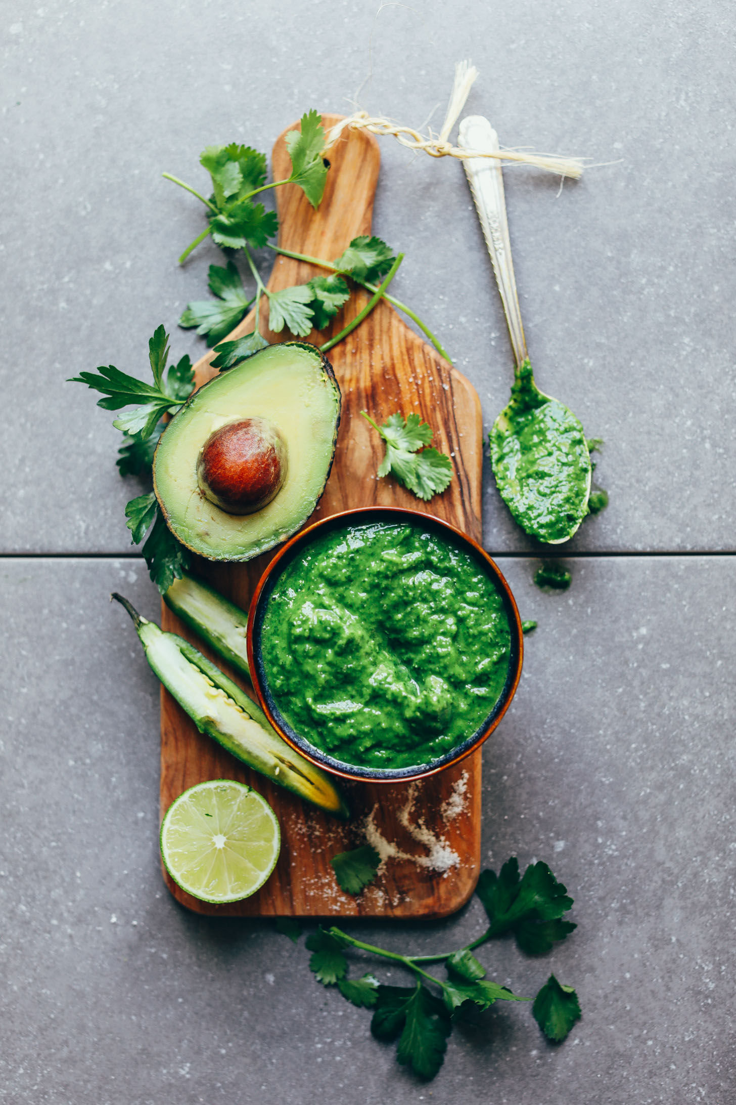 Wood serving board with a bowl of fresh Green Chimichurri and ingredients used to make it