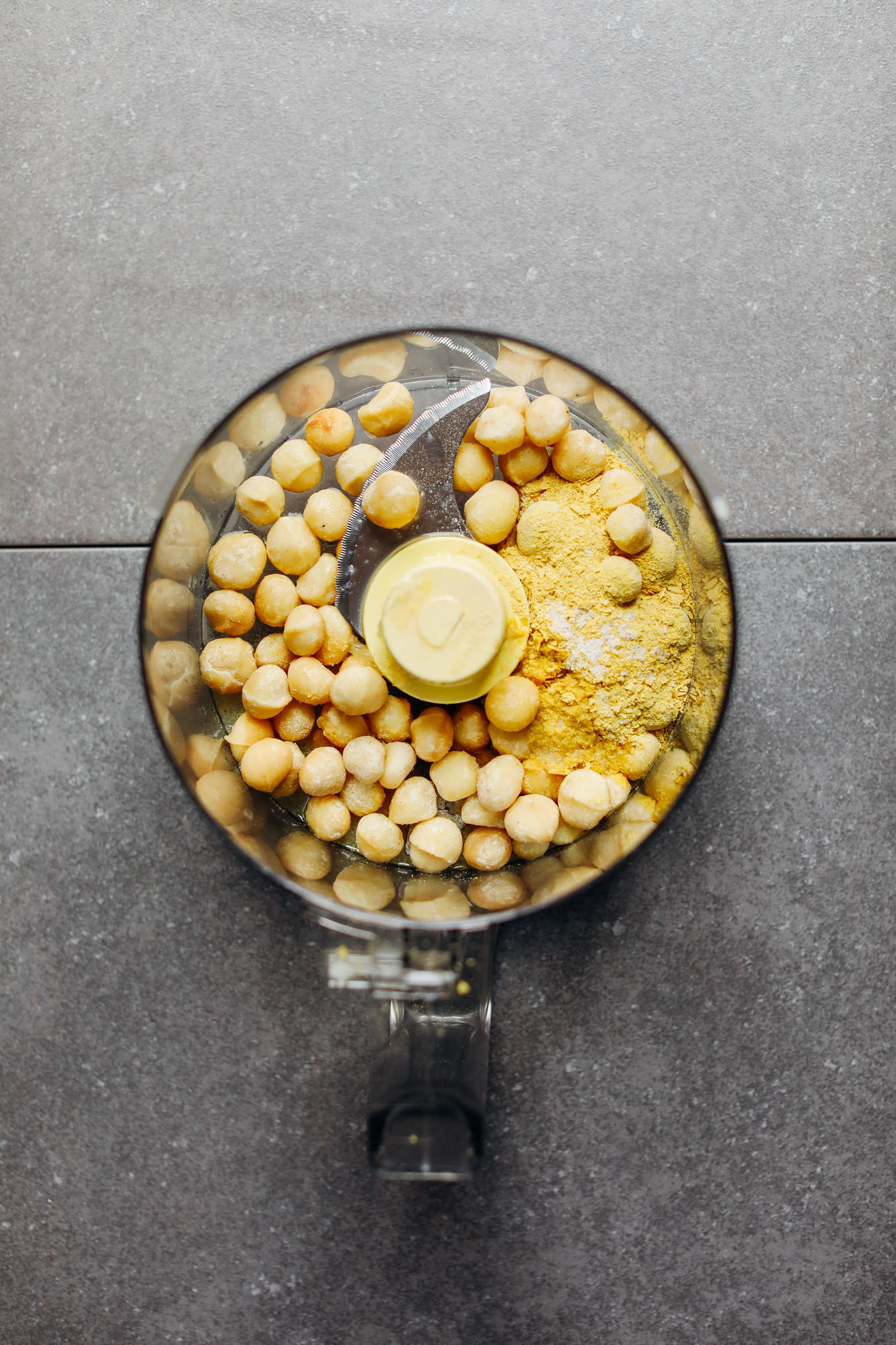Food processor with macadamia nuts, nutritional yeast, garlic powder, and salt for making Macadamia Nut Herb Cheese