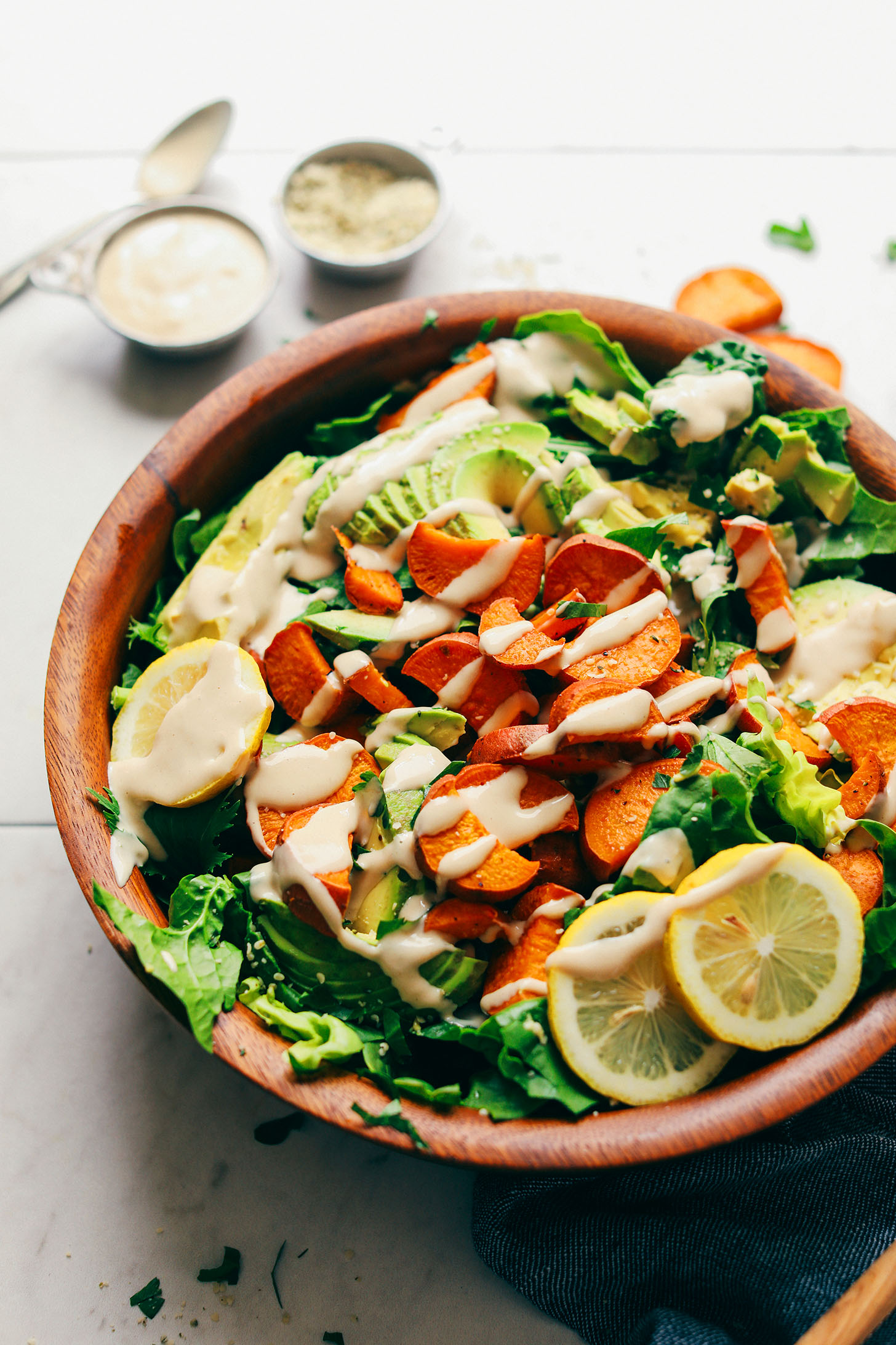 Wood serving bowl with fresh greens, roasted sweet potato, avocado, sliced lemons, and tahini dressing