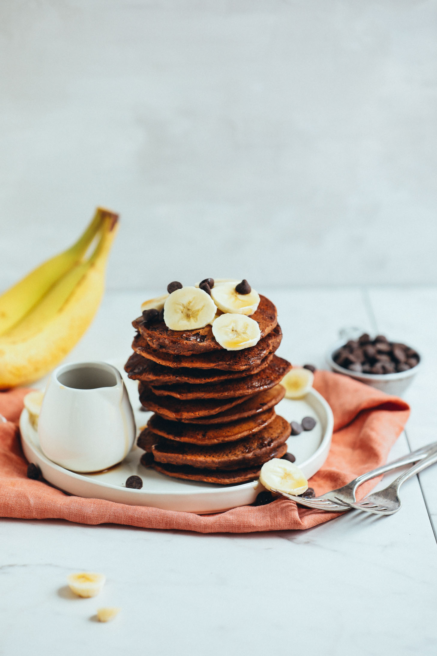 Stack of Vegan Chocolate Chip Pancakes with banana and a side of syrup