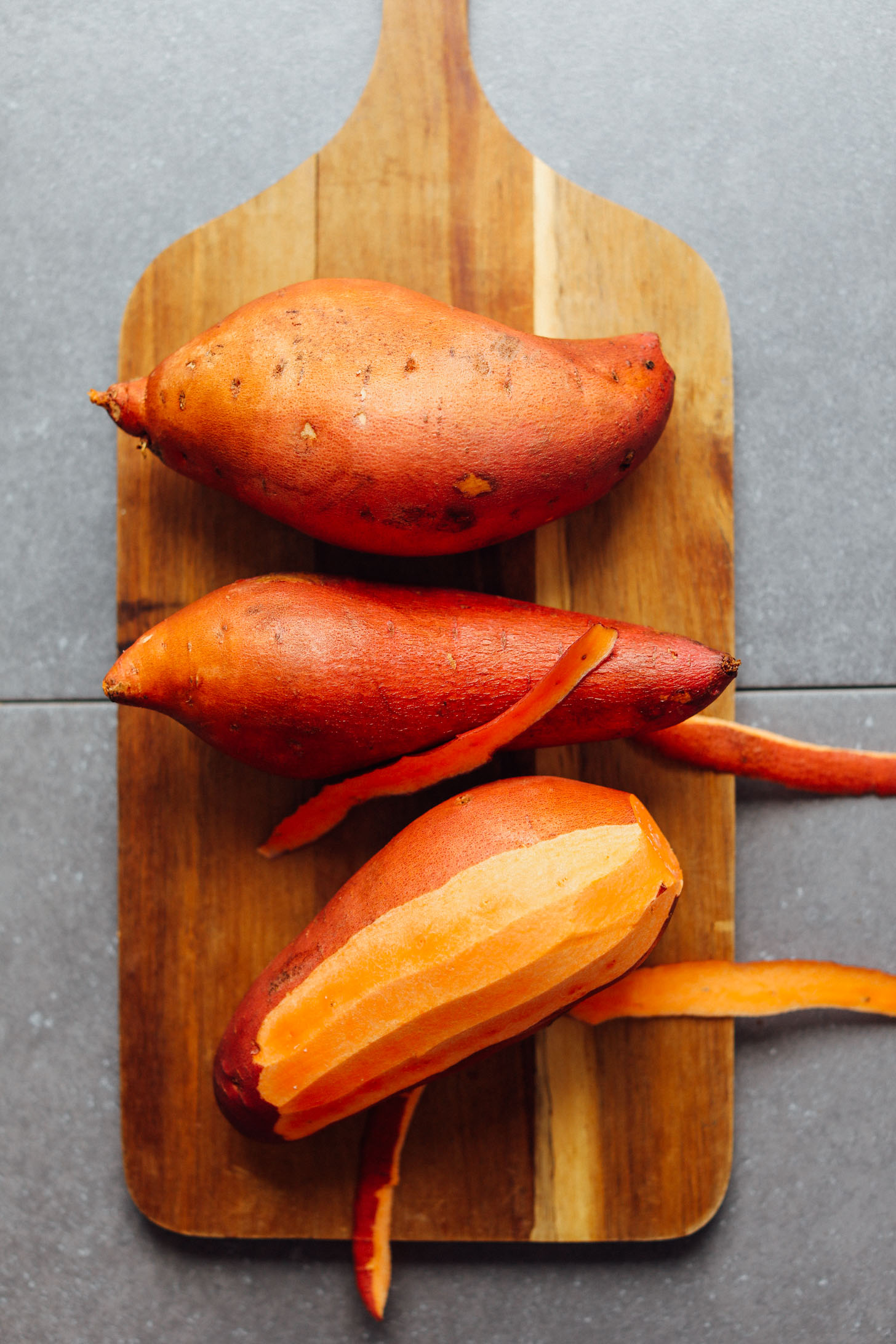 Wood cutting board with sweet potatoes, one of which is partially peeled