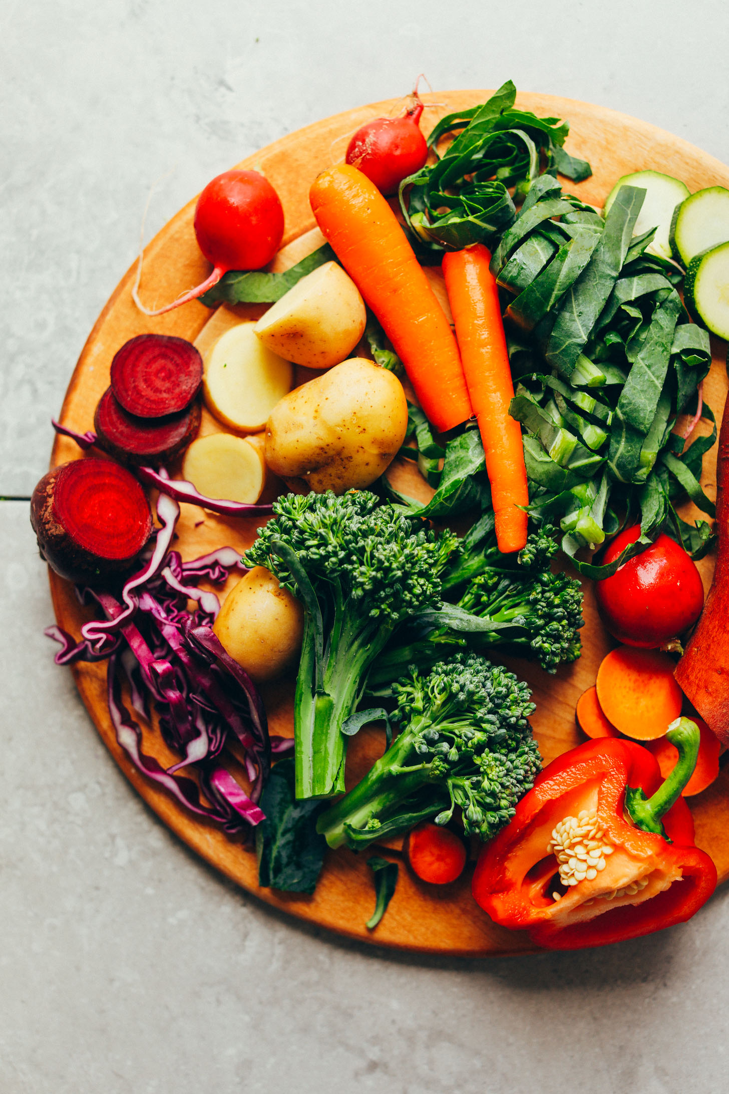 Wood cutting board full of beets, radishes, potatoes, carrots, collard greens, zucchini, sweet potato, broccolini, and red bell pepper for making a delicious plant-based bowl