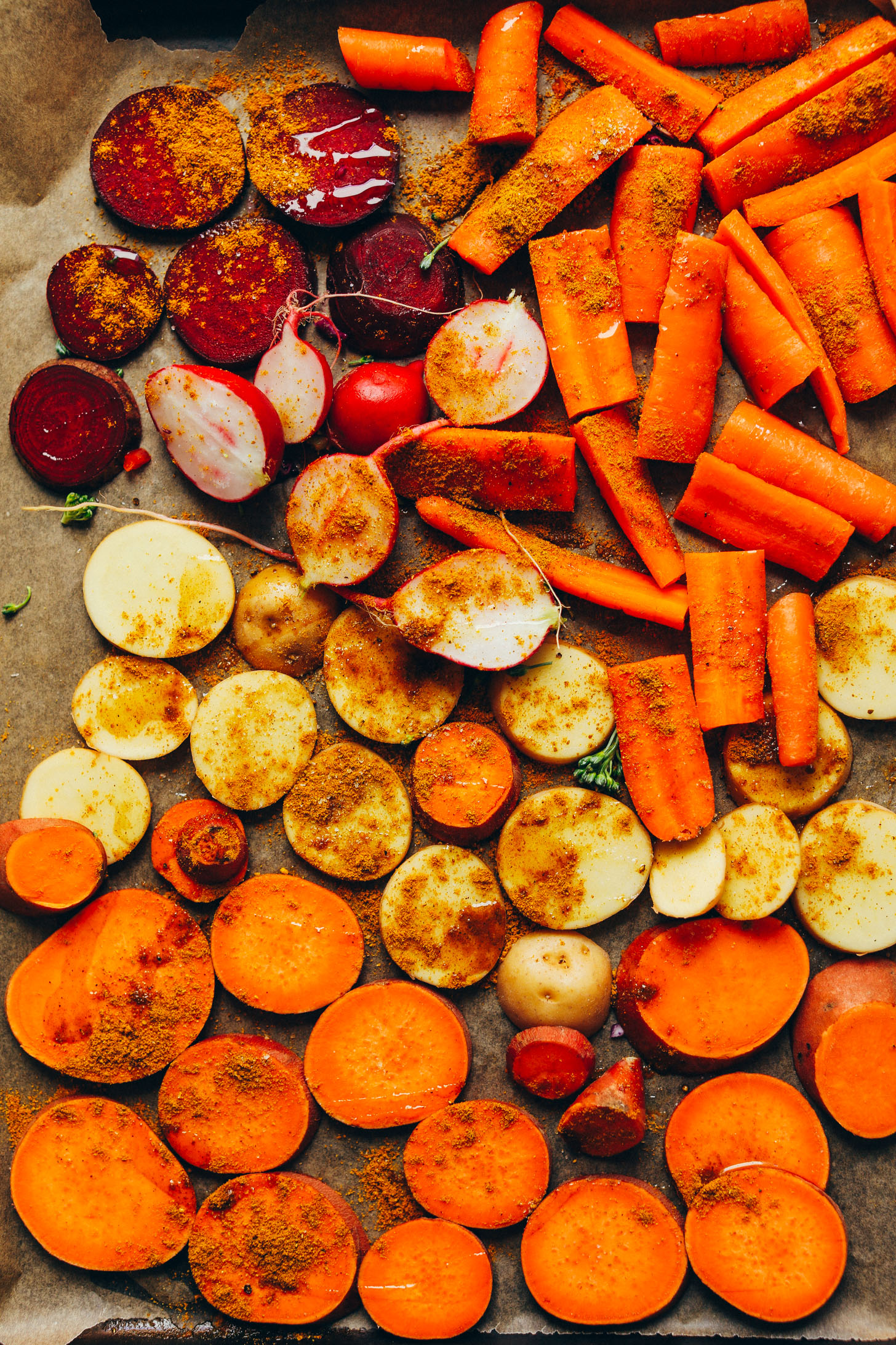 Parchment-lined baking sheet with fresh carrots, radishes, potatoes, sweet potatoes, and beets sprinkled with curry powder