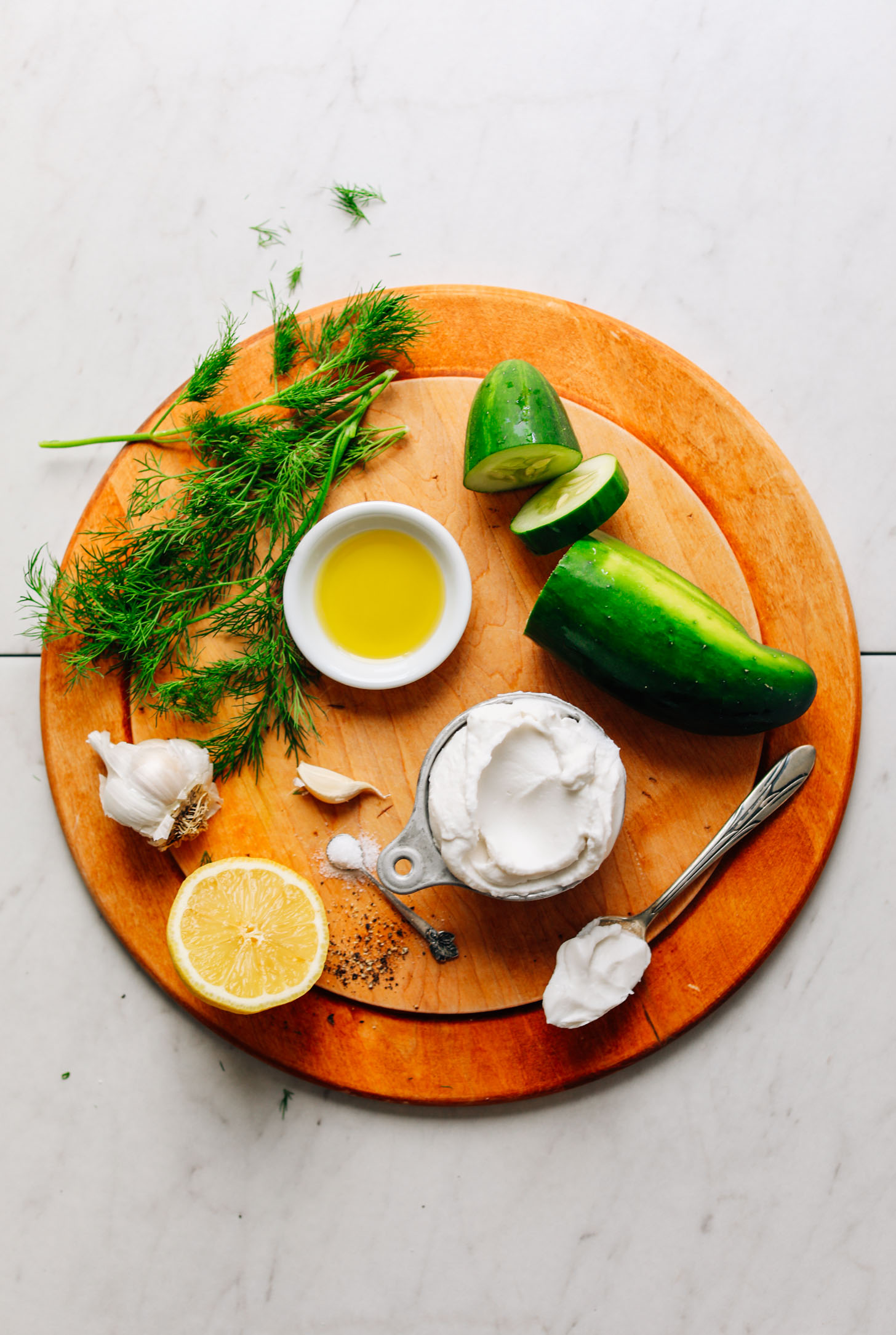 Wood cutting board with coconut yogurt, lemon, garlic, olive oil, cucumber, salt, and fresh dill for making Vegan Tzatziki