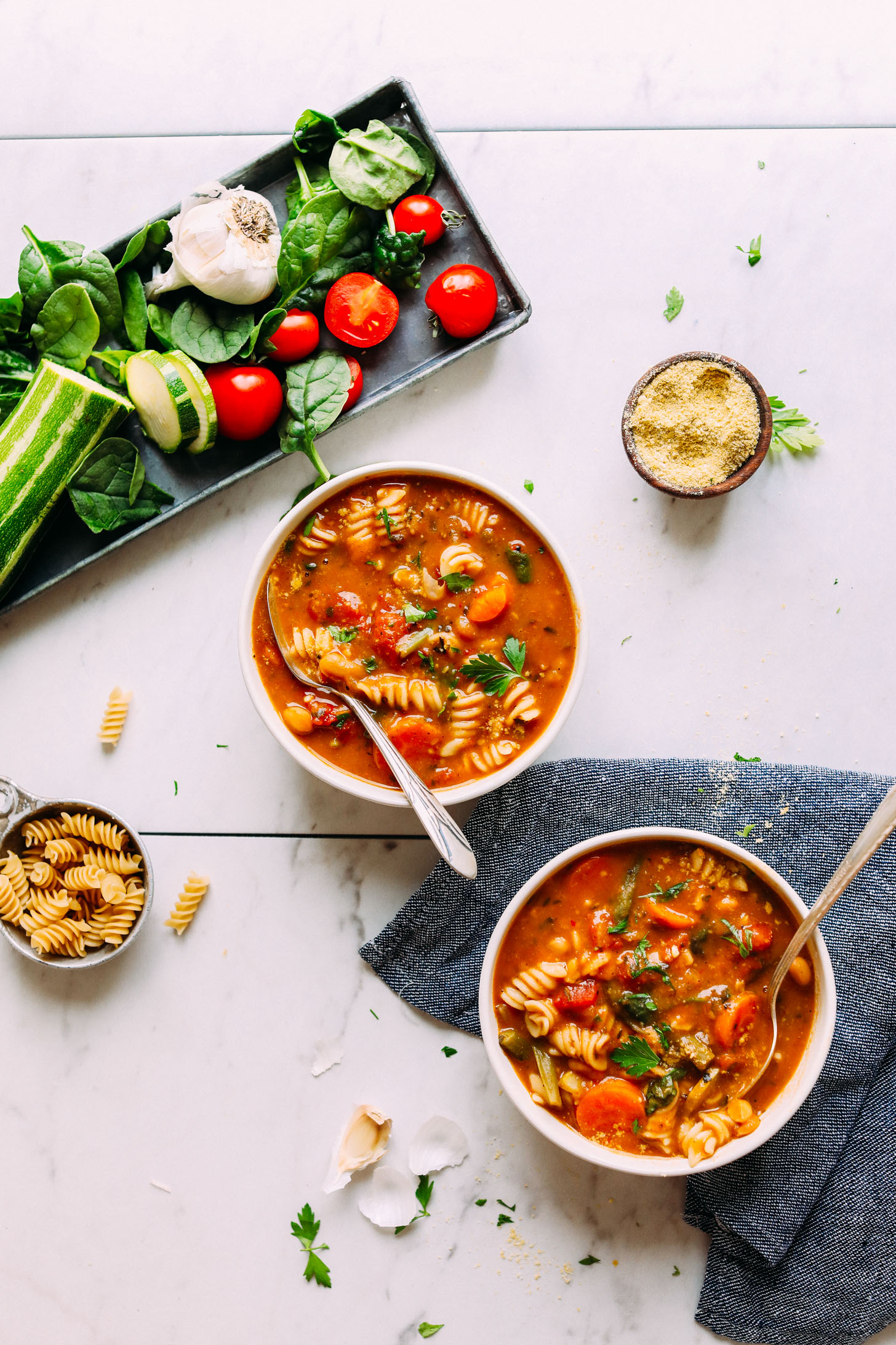 Two bowls of 1-Pot Vegan Minestrone alongside fresh vegetables used to create it