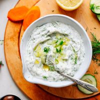 Bowl of homemade Vegan Tzatziki beside carrots, cucumber, dill, and lemon