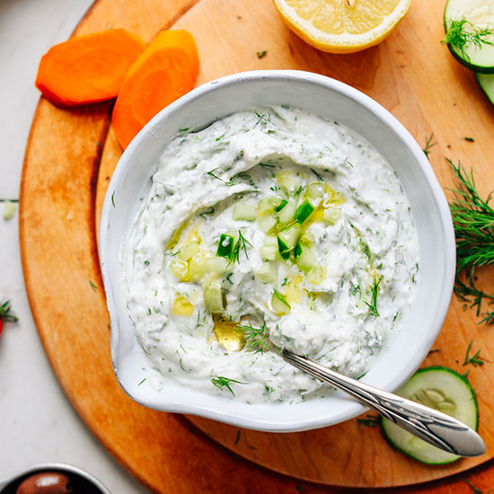 Bowl of homemade Vegan Tzatziki beside carrots, cucumber, dill, and lemon