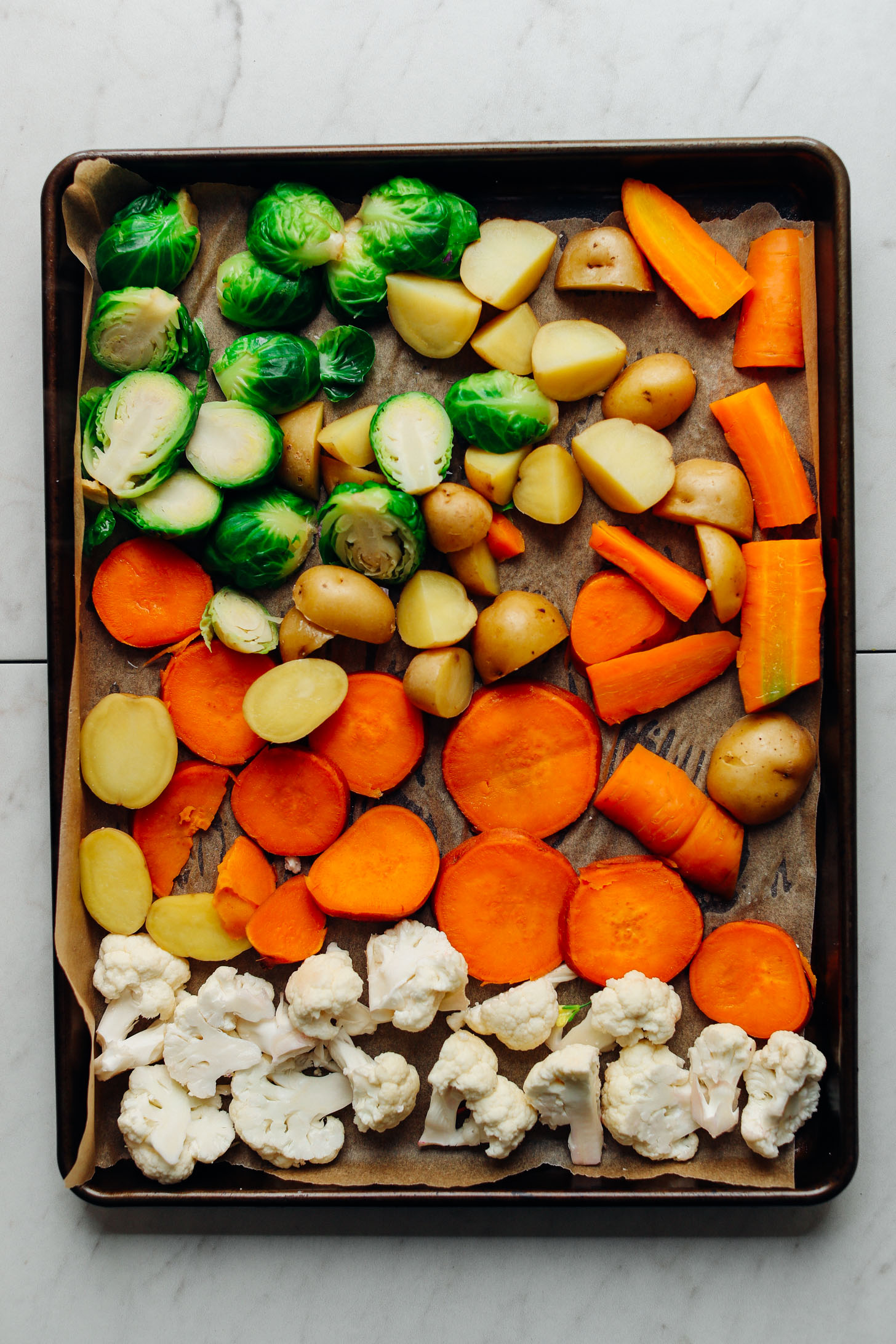 Baking sheet of chopped cauliflower, sweet potatoes, carrots, potatoes, and Brussels Sprouts for our oven roasted vegetables recipe