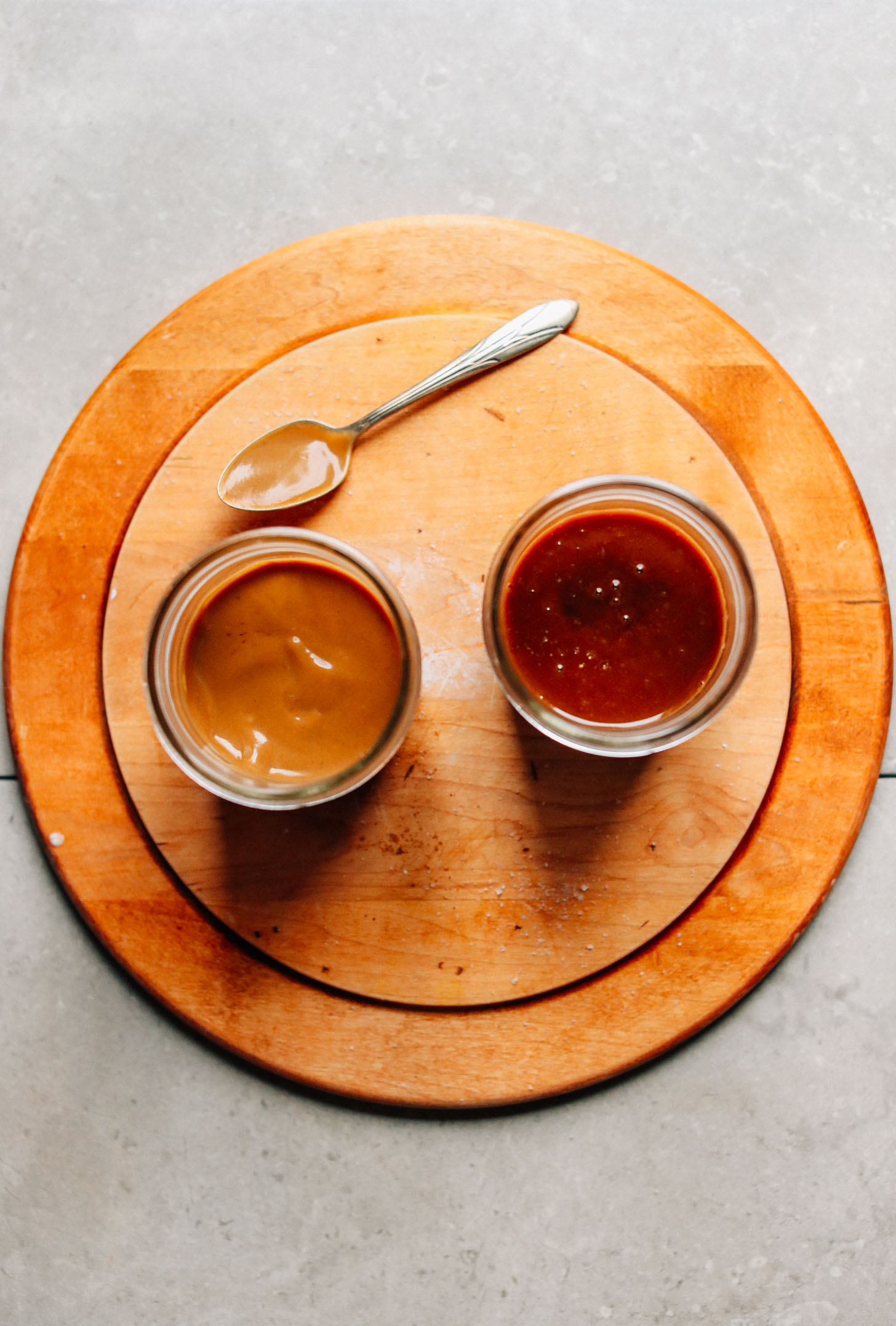 Two jars of refined-sugar-free Vegan Caramel Sauce on a wood cutting board with a spoon