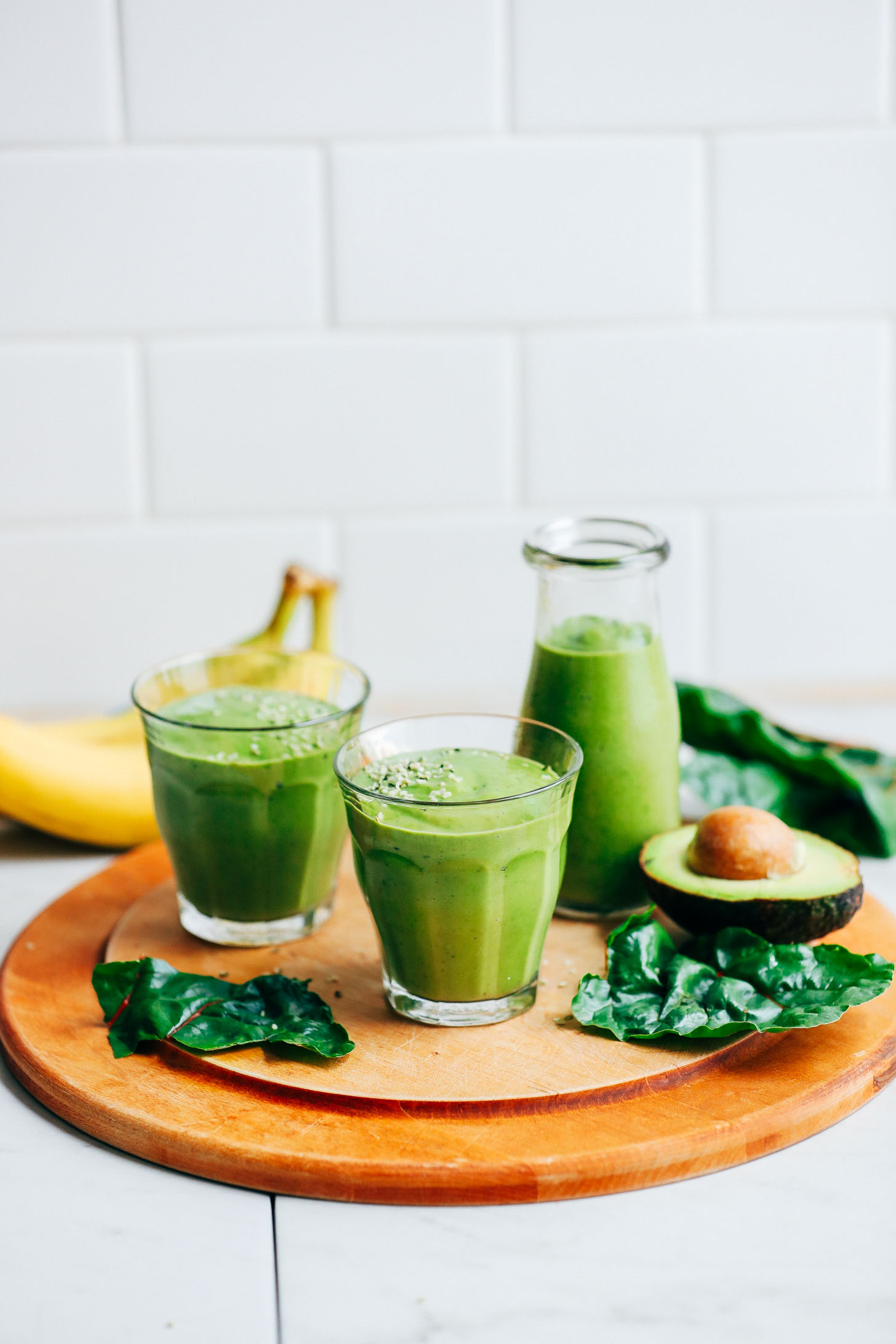 Two serving glasses filled with vibrant Avocado Green Smoothie and surrounded by avocado, banana, and kale