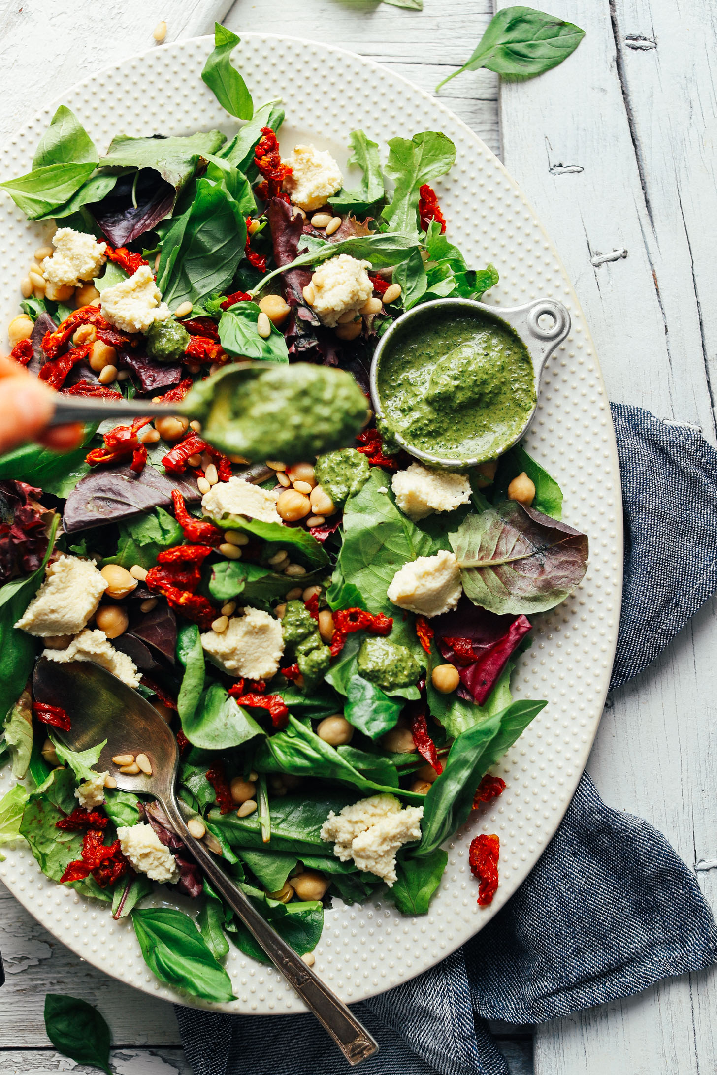 Using a spoon to drizzle Vibrant Pesto over salad with almond ricotta and sun-dried tomatoes