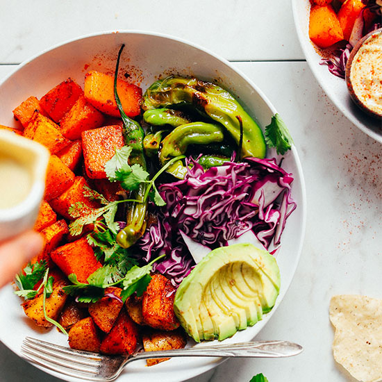 Pouring a small jar of Vegan Jalapeño Queso onto a bowl of roasted butternut squash and other veggies