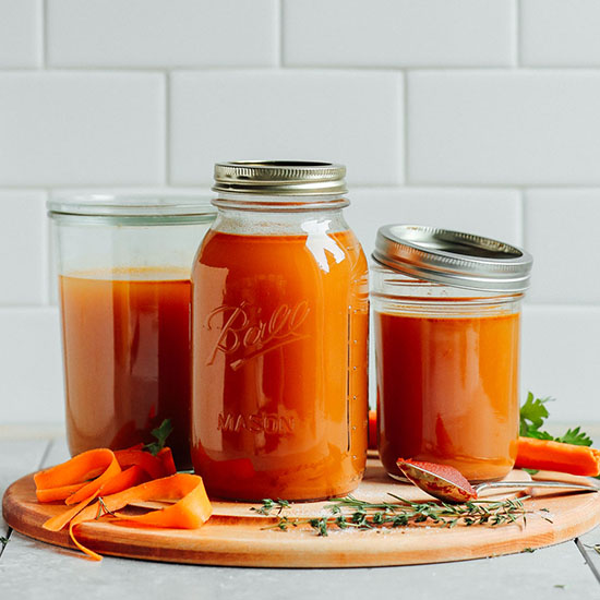 Jars of homemade Easy 1-Pot Vegetable Broth on a cutting board with tomato paste, carrots, and herbs