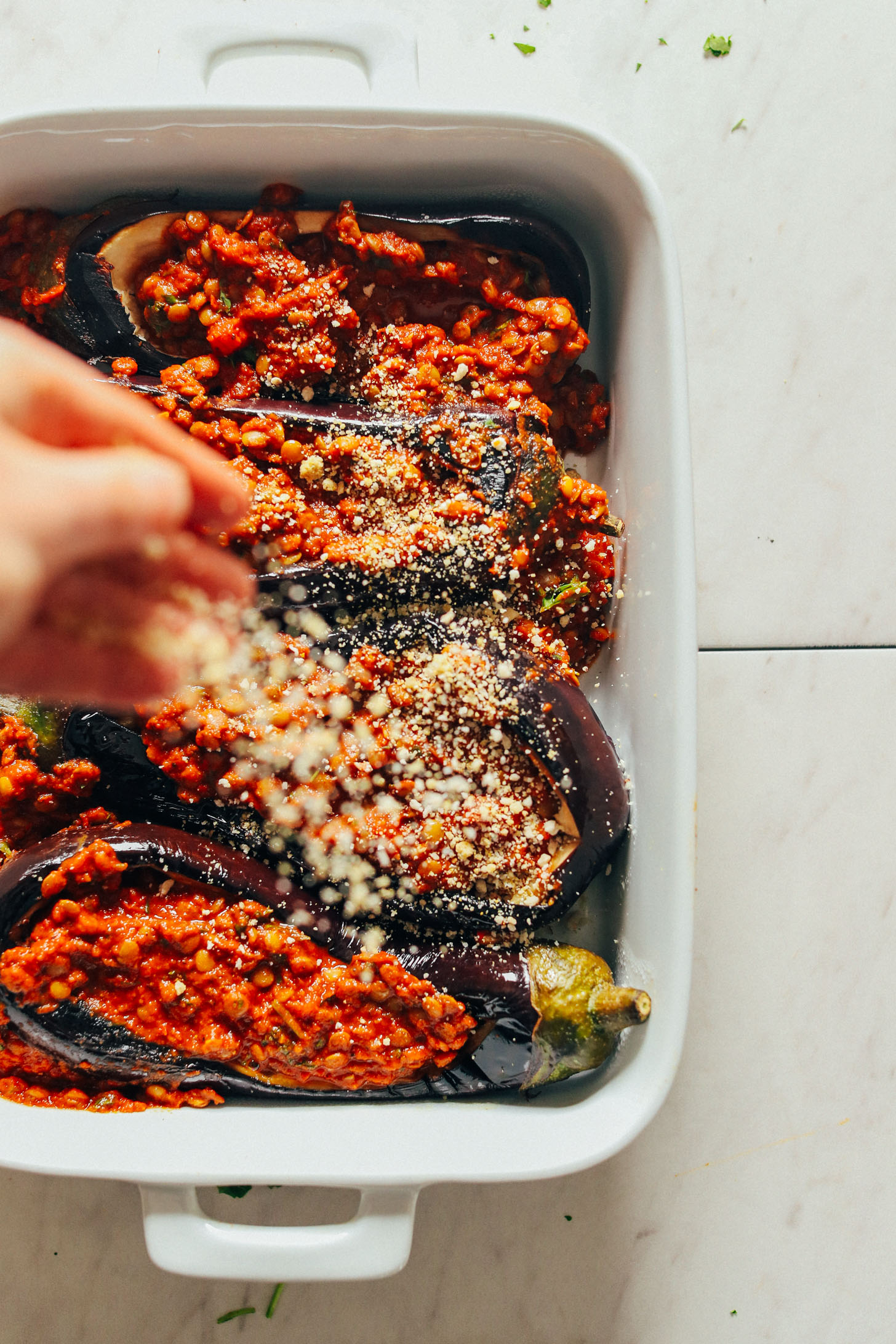 Sprinkling vegan parmesan cheese over a ceramic baking dish full of Moroccan Lentil-Stuffed Eggplant