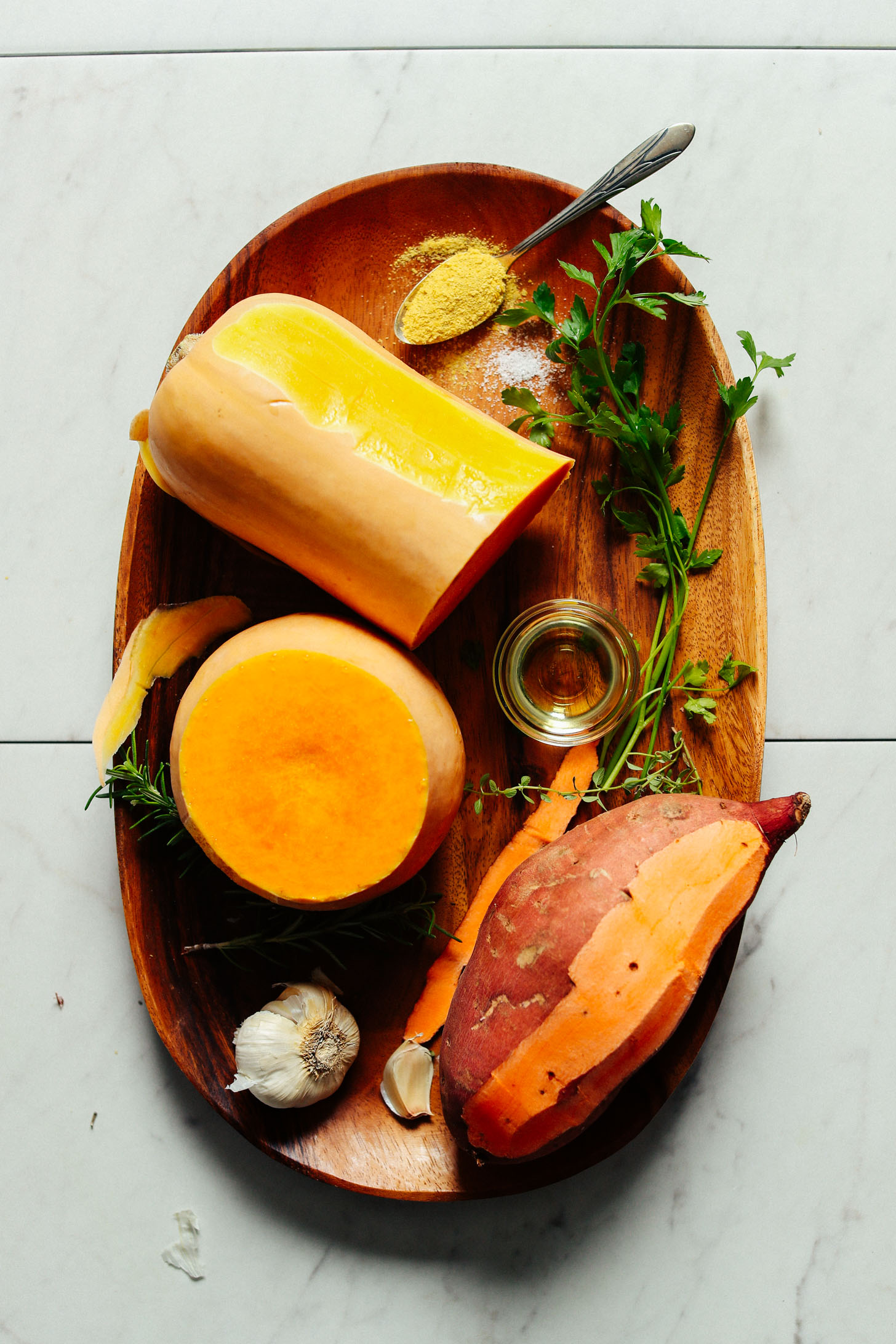Butternut squash, sweet potato, garlic, olive oil, parsley and nutritional yeast displayed on a wooden platter