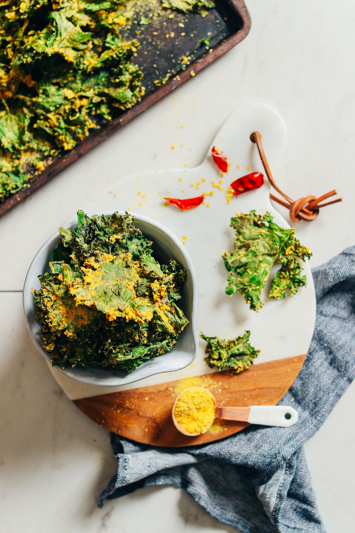 Marble cutting board with a bowl of Crispy Baked Kale Chips coated with Sunflower Seed Cheeze