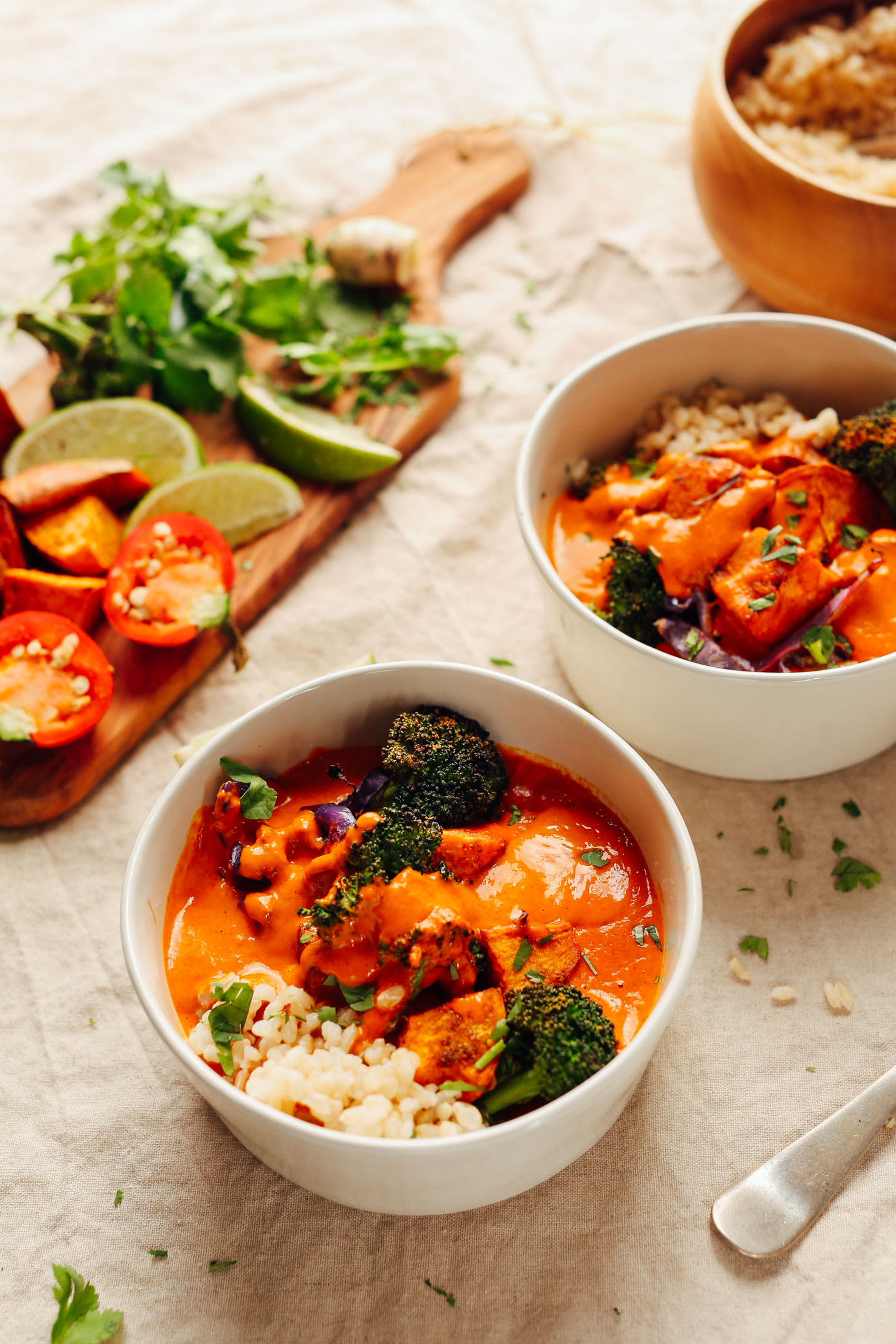 Bowls of Rich Red Curry with Roasted Vegetables alongside a cutting board with fresh limes, peppers, and herbs for serving
