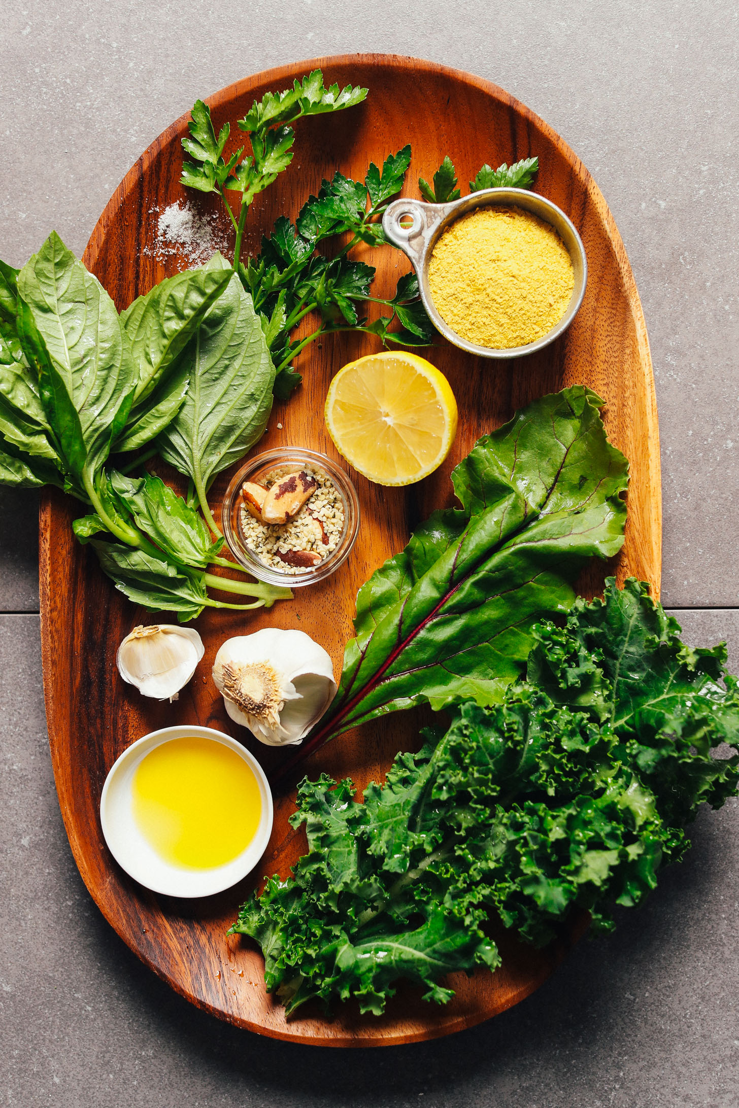A platter full of ingredients for making Super-Green Vegan Pesto- including olive oil, garlic, kale, beet greens, basil, Brazil nuts, hemp seeds, lemon, parsley, and nutritional yeast