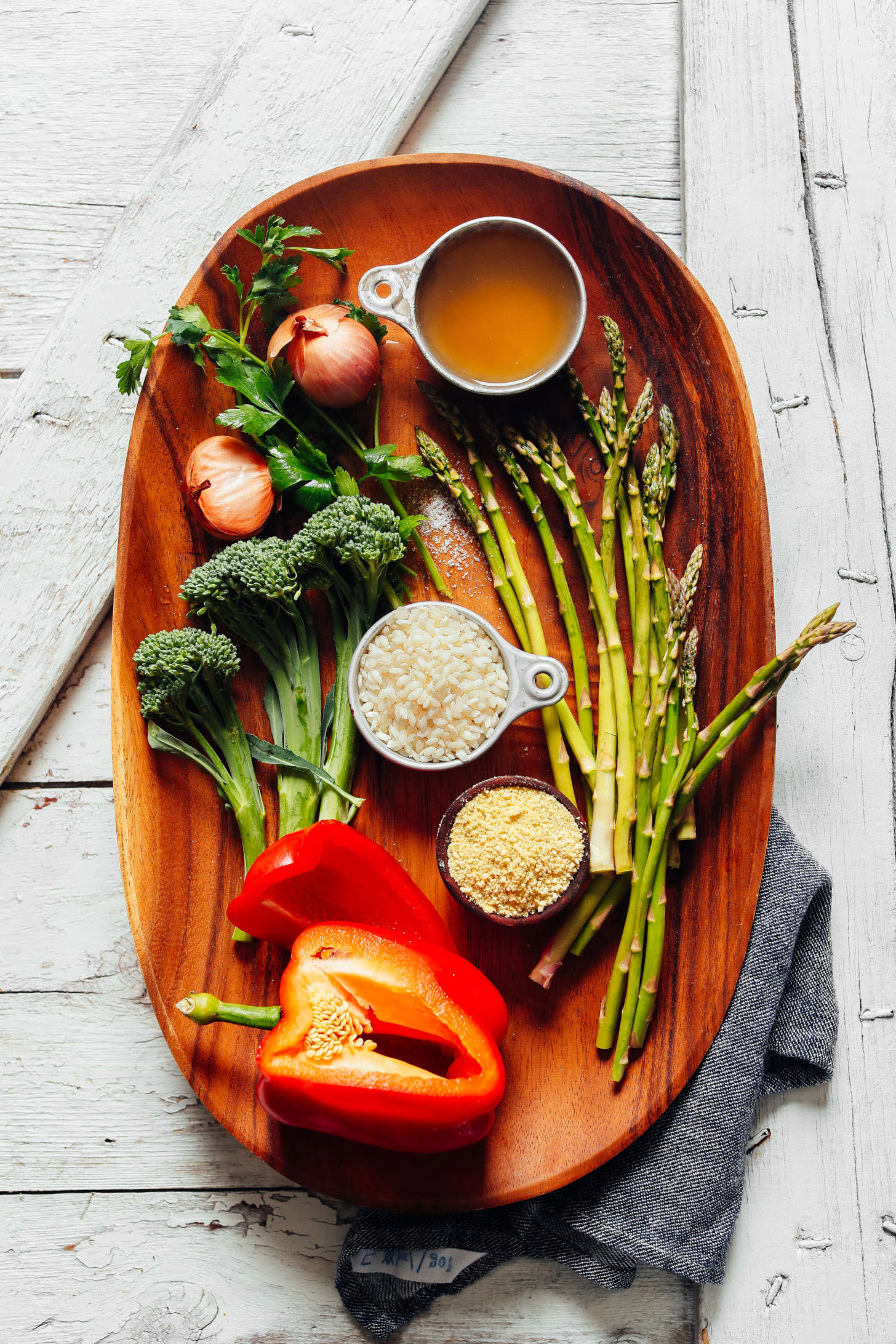 Wood serving platter displaying broccolini, shallots, parsley, red bell pepper, arborio rice, asparagus, vegetable broth, and vegan parmesan for making Creamy Vegetable Risotto