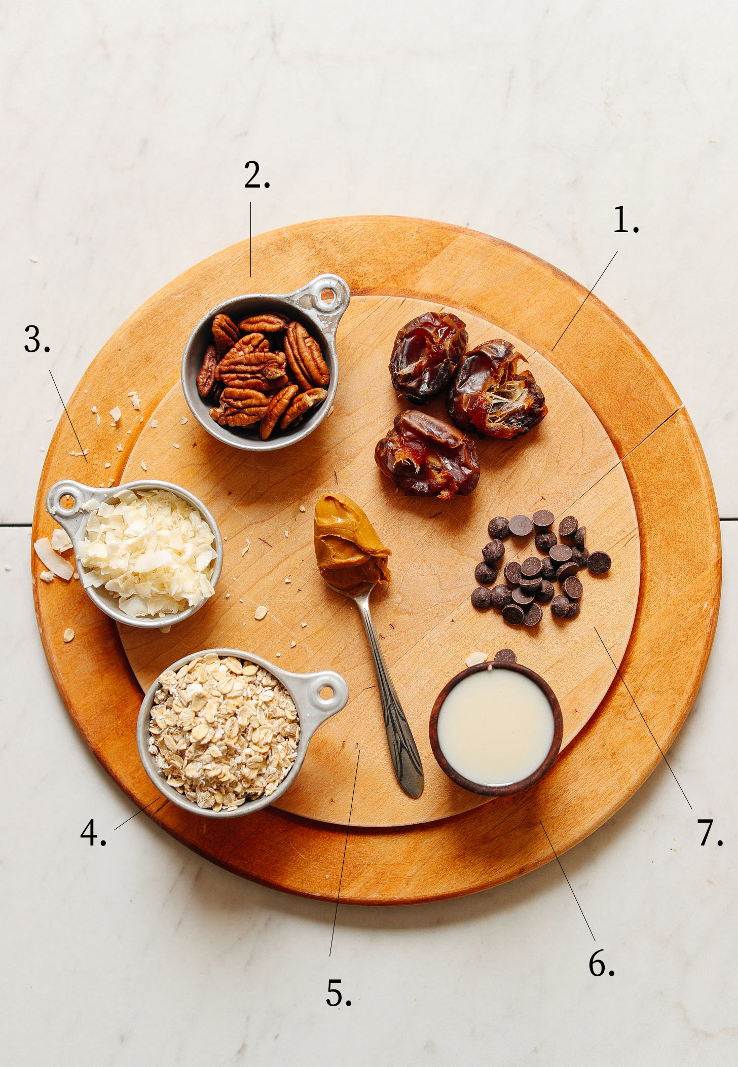 Wooden cutting board displaying dates, pecans, coconut flakes, rolled oats, peanut butter, coconut butter and vegan chocolate chips for making Vegan 7-Layer Bars