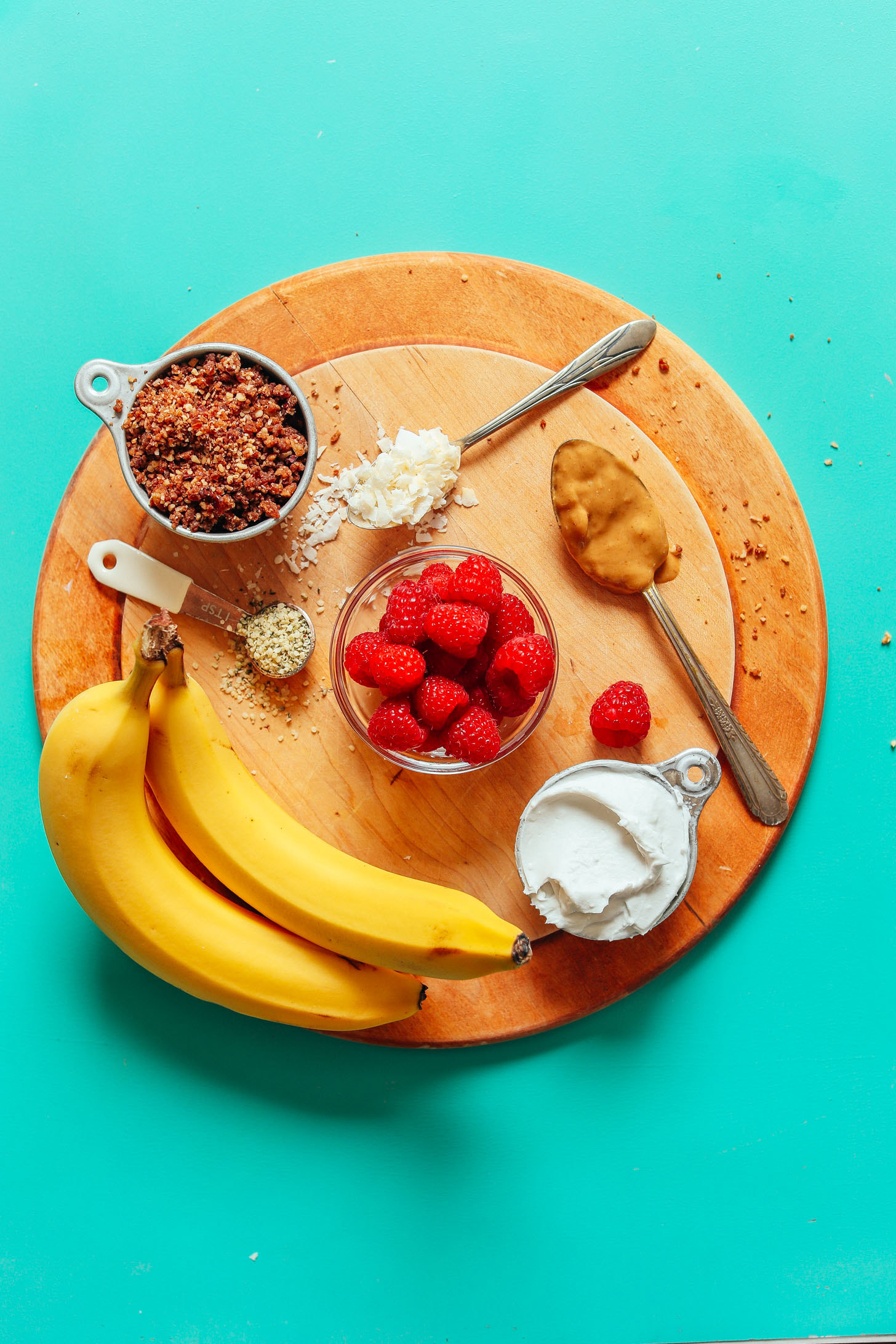 Display of ingredients for Raw Banana Splits including bananas, raspberries, coconut yogurt, flaked coconut, peanut butter, and rawnola