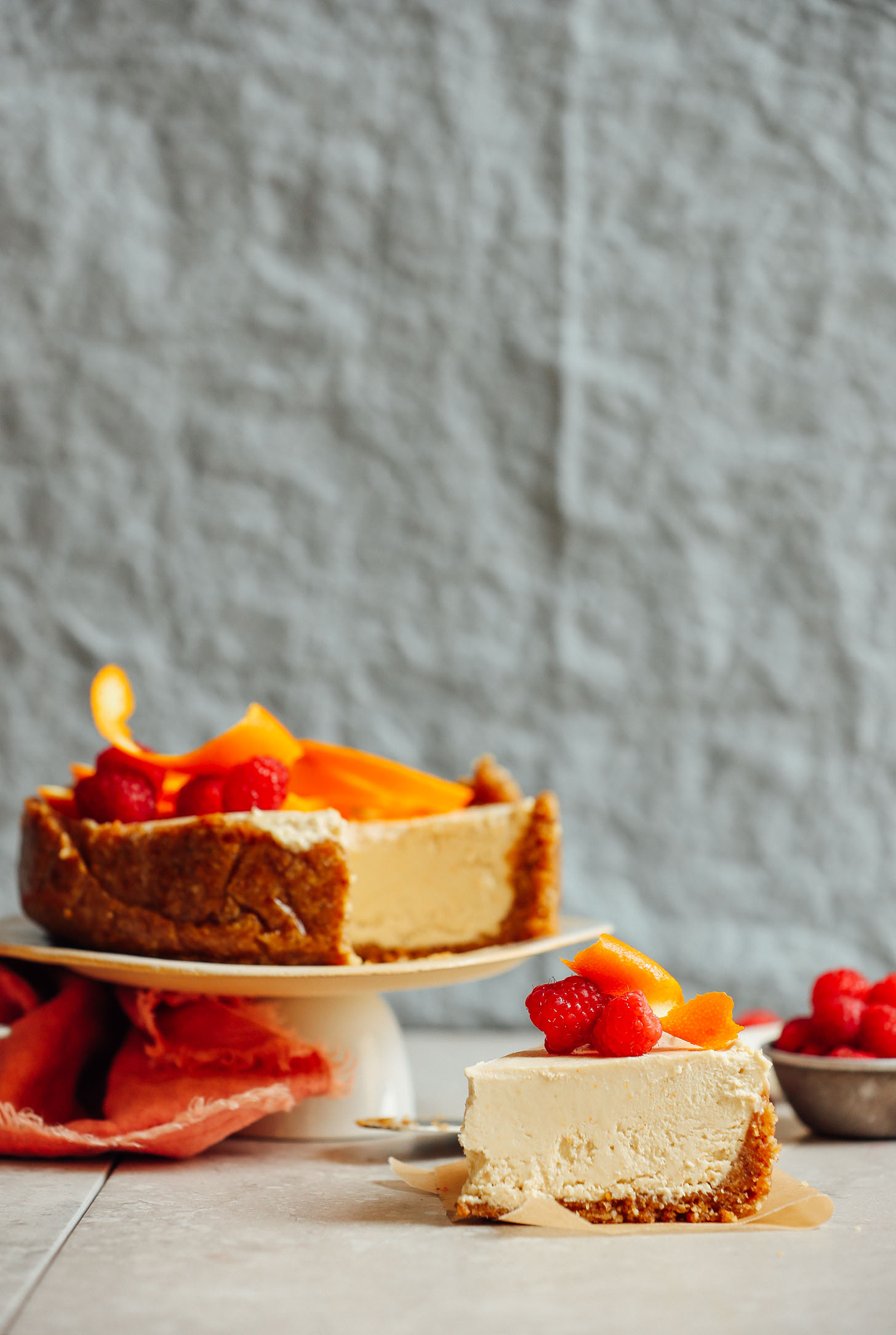 Slice of vegan gluten-free Coconut Yogurt Cheesecake in front of the remainder of the dessert that is perched on a serving platter