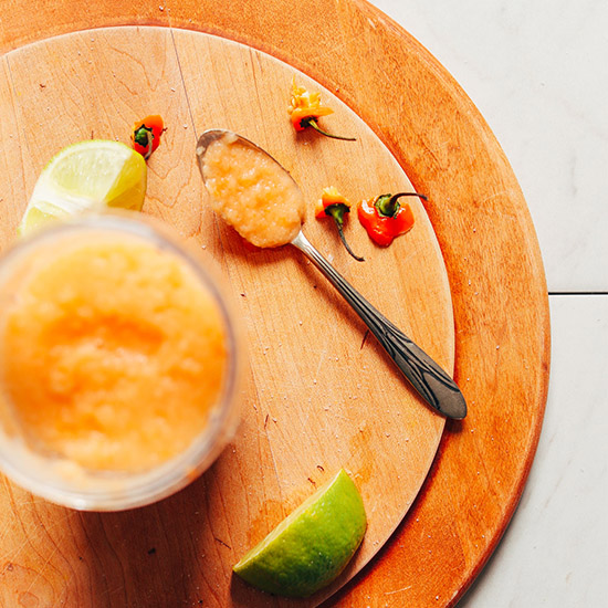 Spoon and container of homemade Habanero Hot Sauce beside lime wedges and habanero stems
