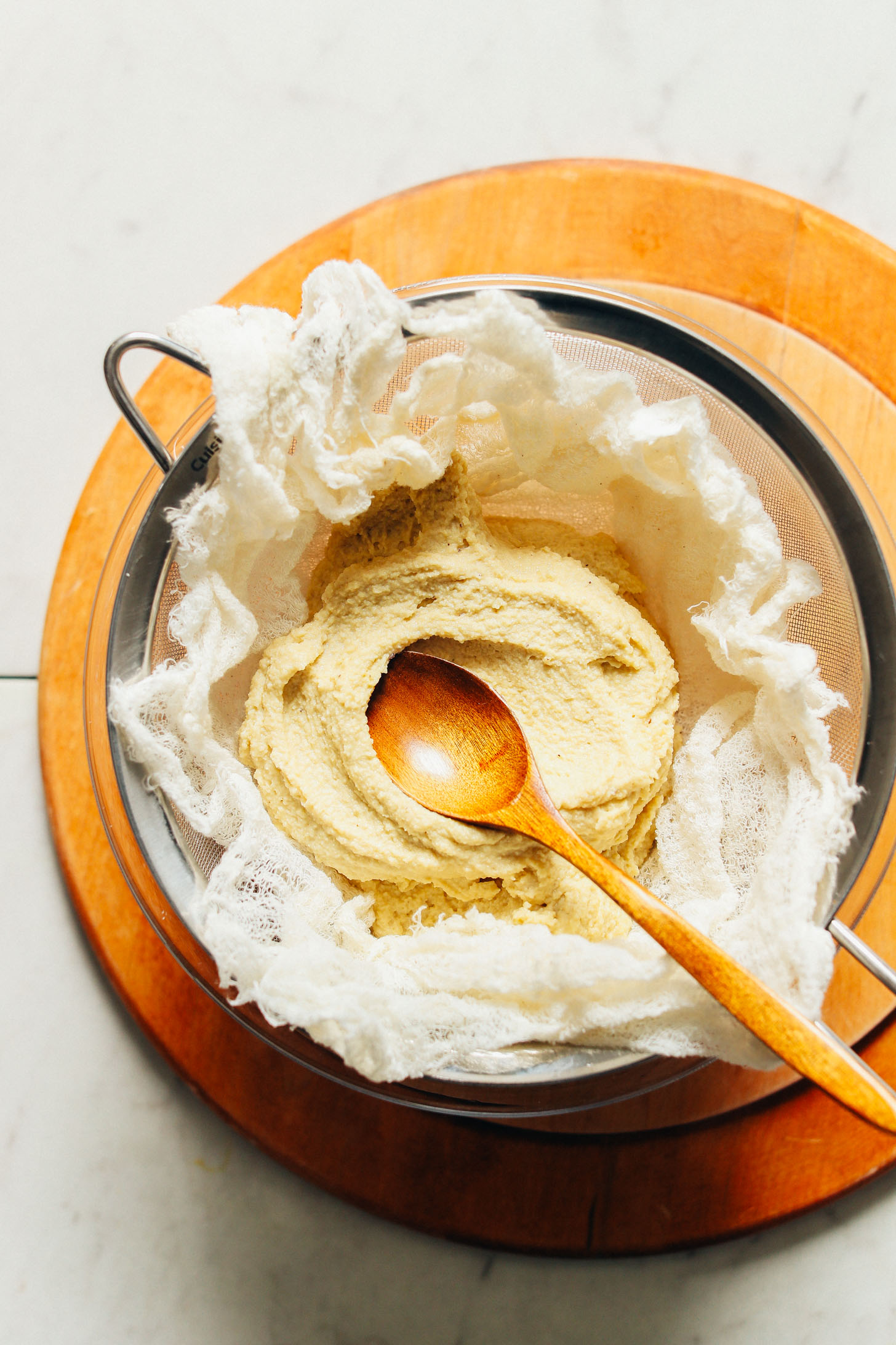 Using a wooden spoon to flatten out homemade vegan cheese in a cheesecloth