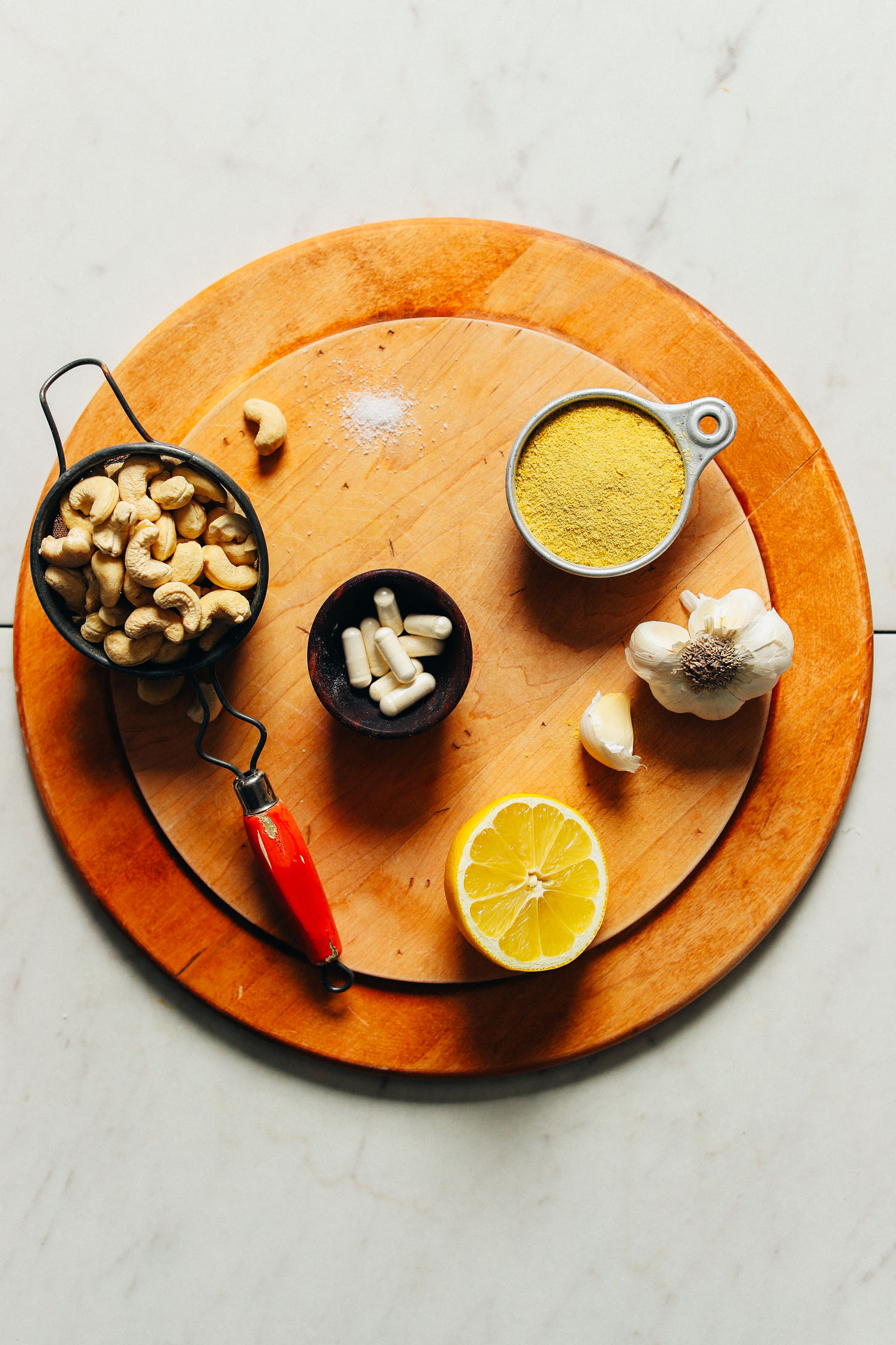 Wood cutting board displaying cashews, nutritional yeast, garlic, lemon, salt, and probiotics for homemade vegan cheese