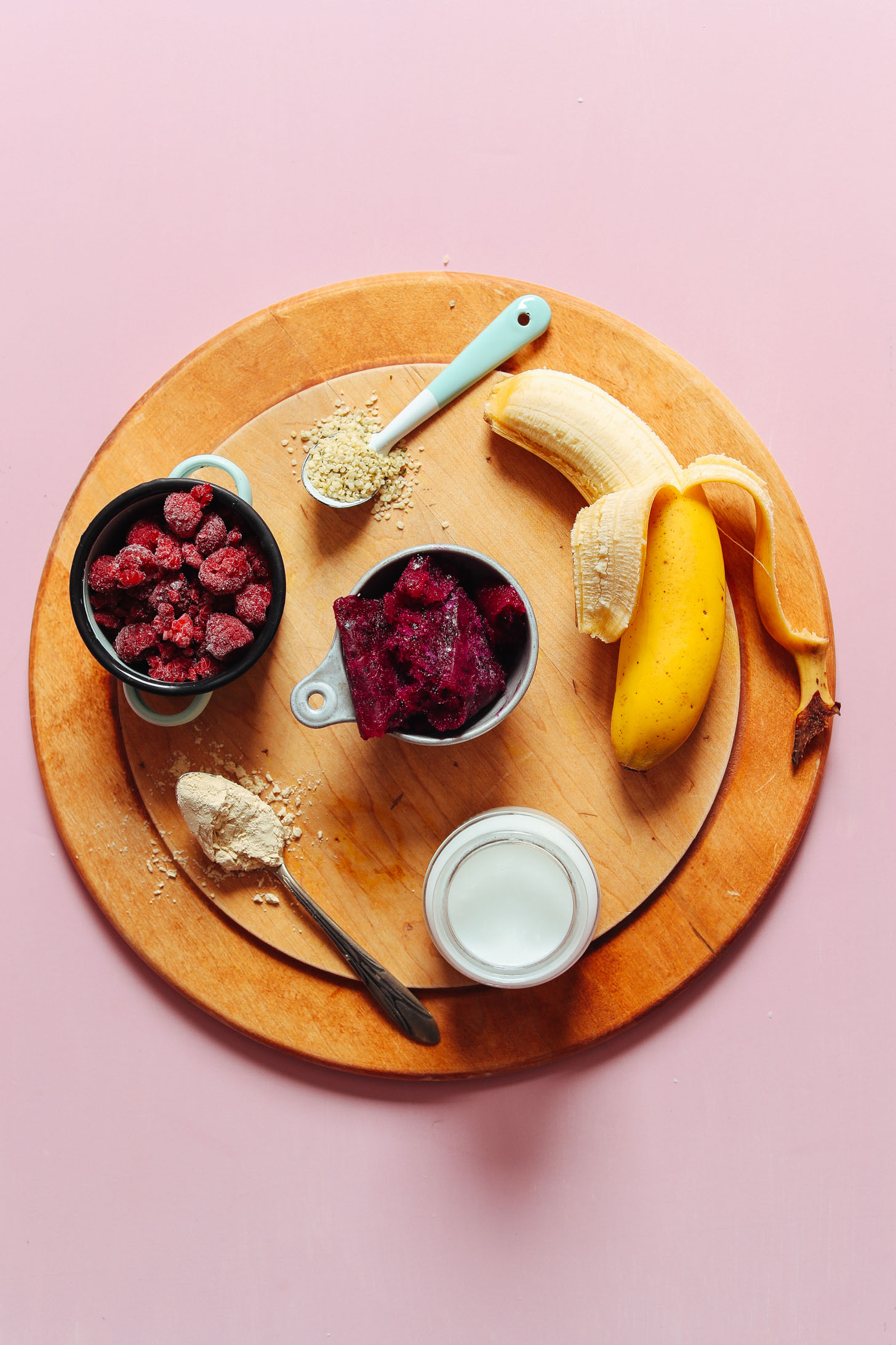 Wood cutting board with banana, hemp seeds, frozen raspberries, dragonfruit, vegan protein powder, and coconut milk for making a Dragon Fruit Smoothie Bowl
