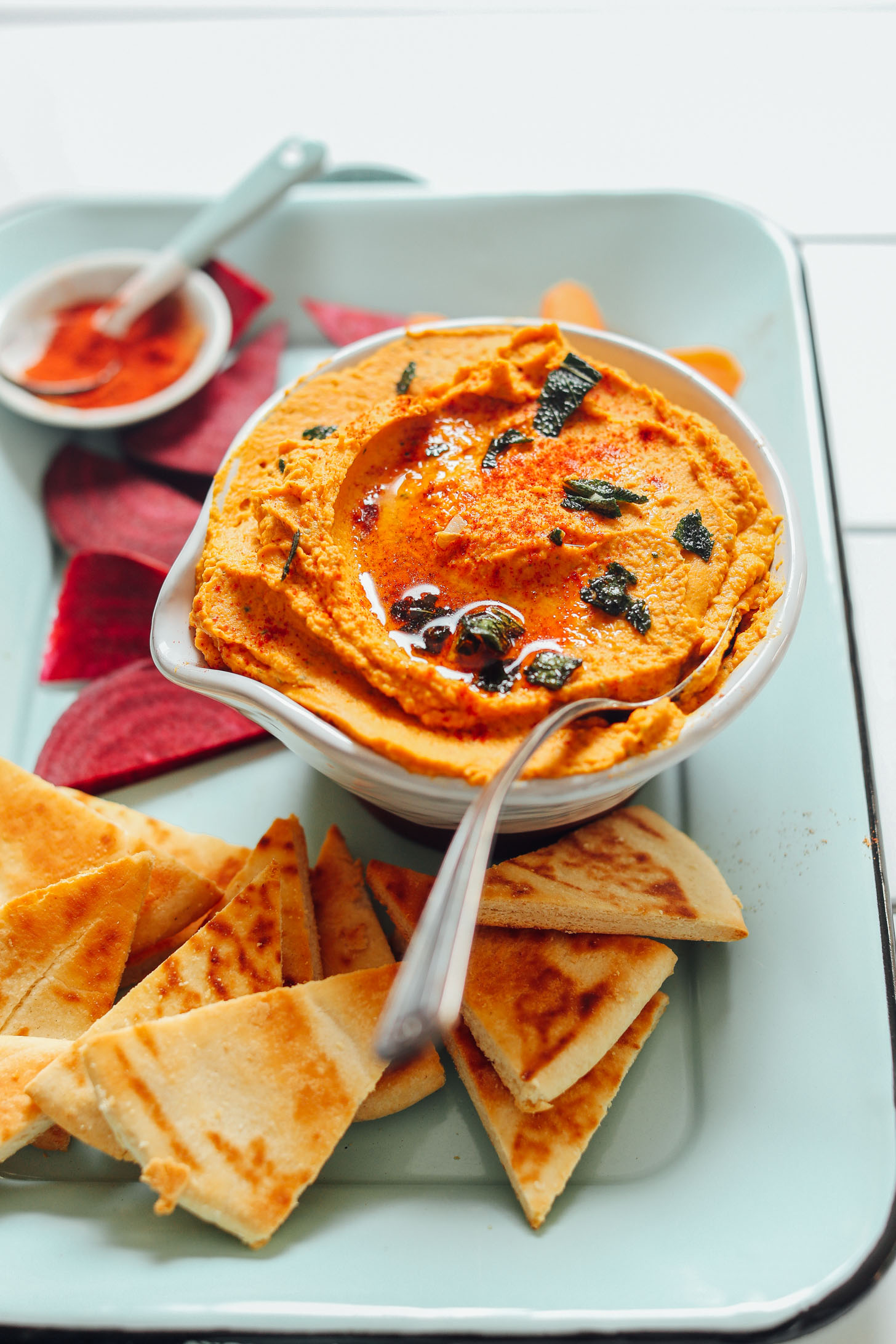 Close up shot of a bowl of White Bean Pumpkin Hummus on a tray with pita slices, beet, and carrot