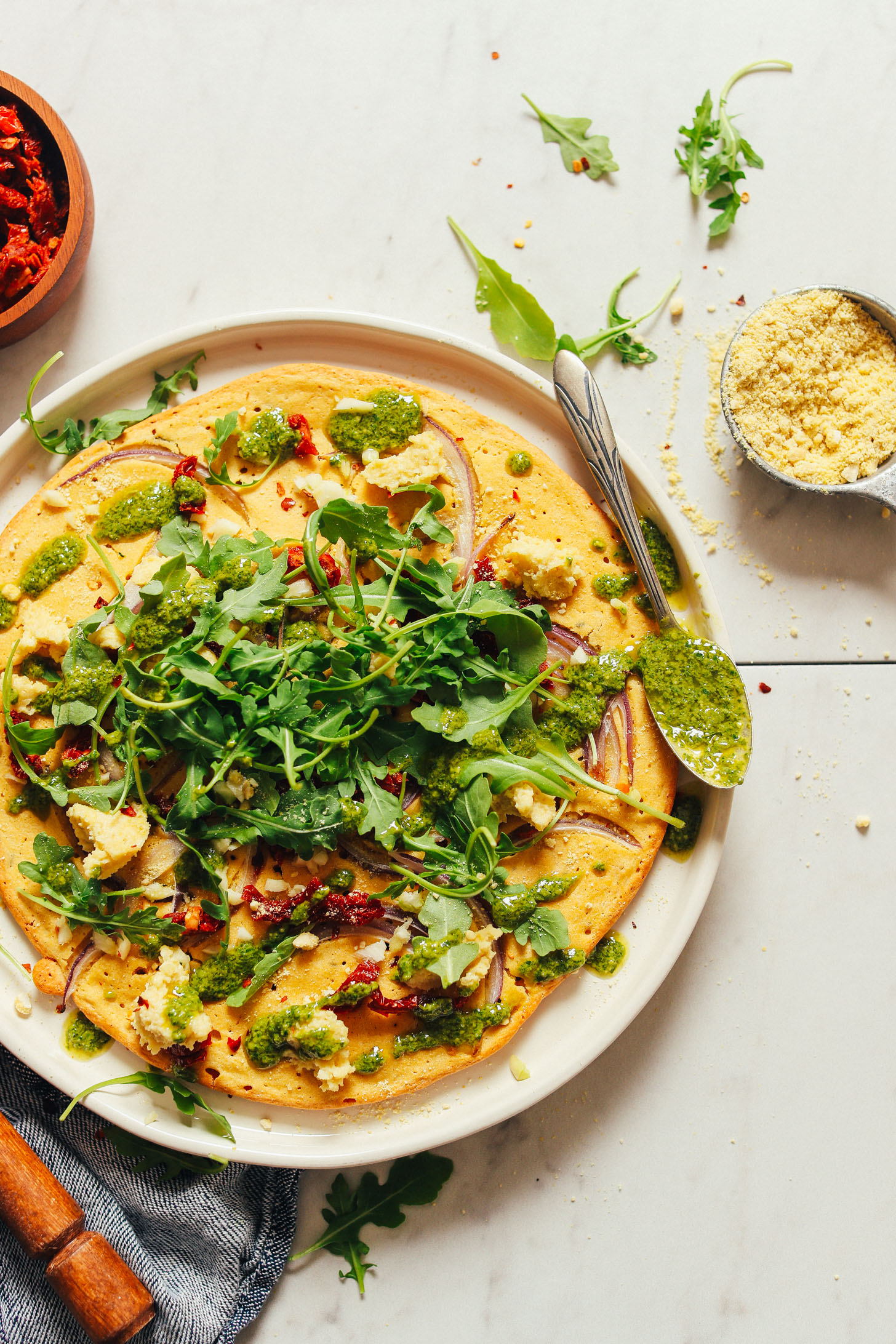 Plate filled with a beautiful gluten-free vegan Socca Pizza with pesto, sun-dried tomatoes, and arugula