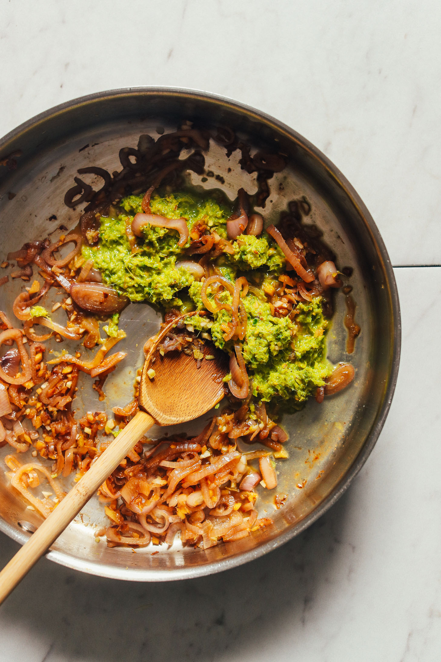 Overhead image of a pan with sautéed onion, garlic, and green curry paste with a wooden spoon stirring the ingredients