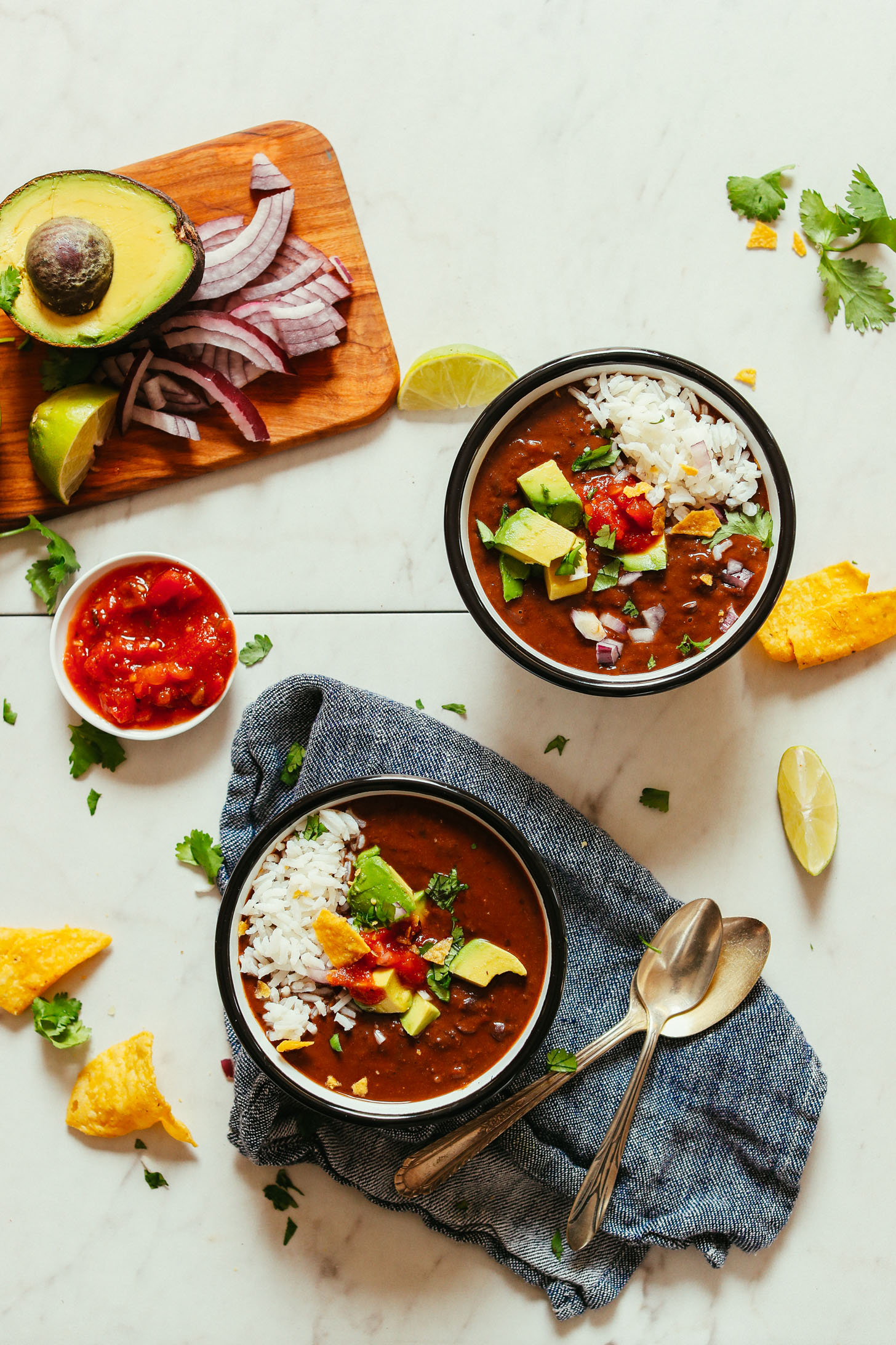 Overhead image of two bowls of black bean soup, one on a blue towel with two spoons, and small cutting board in the top left with ingredients