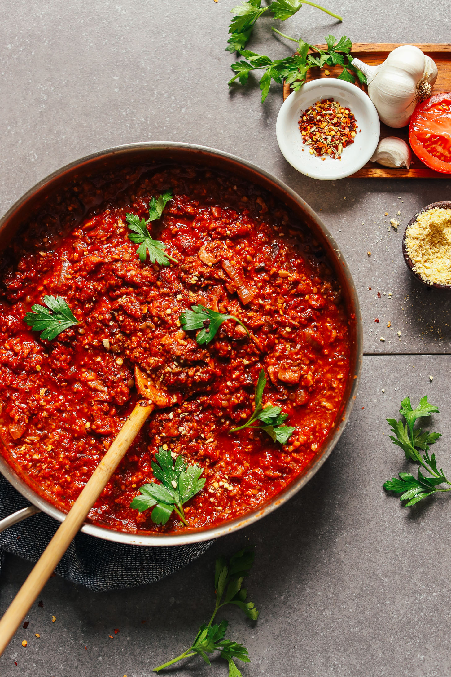 Overhead image of tempeh bolognese in a pan with ingredients in the top right corner, wooden spoon resting in the pan, and parsley as a garnish